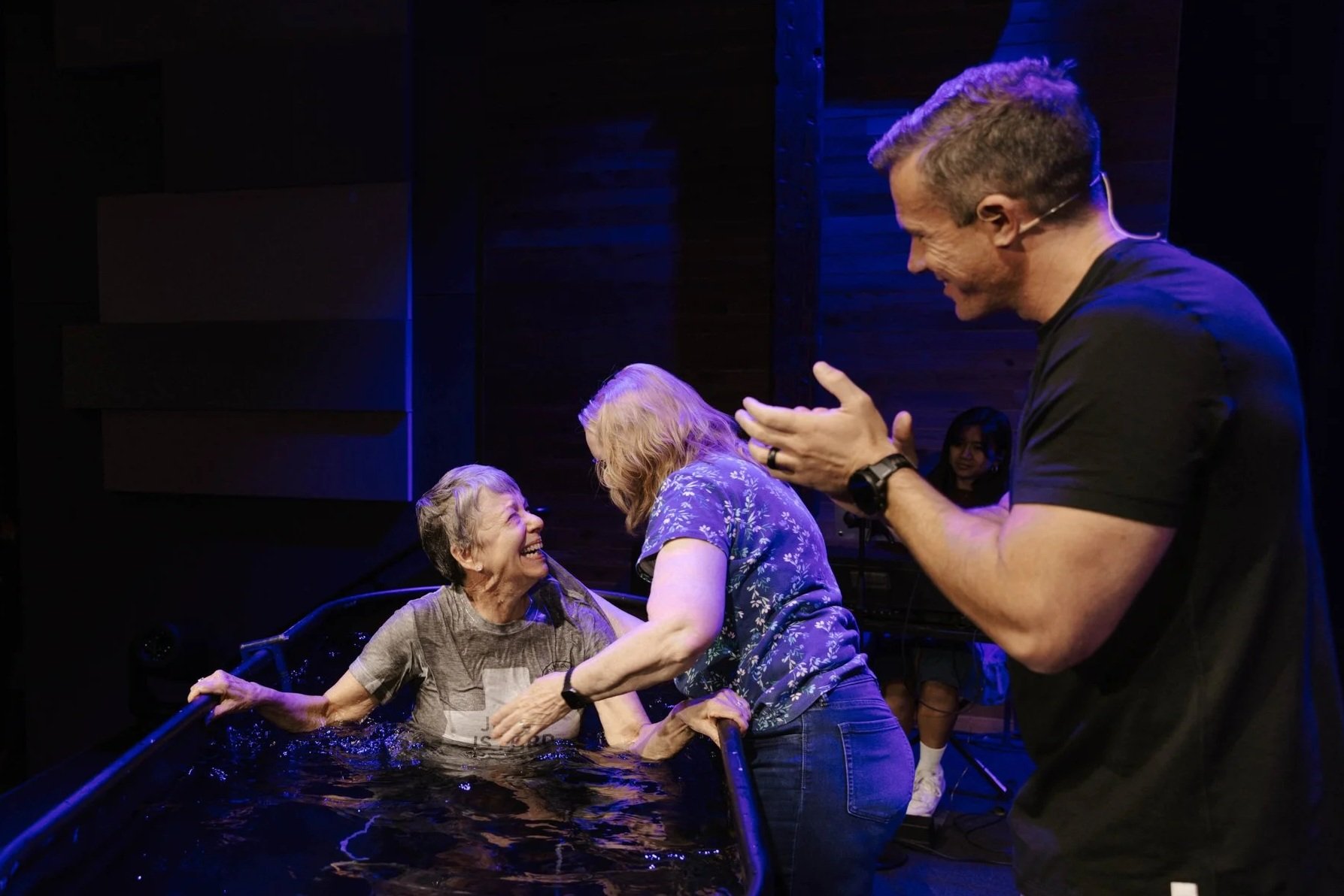 An elderly woman in a baptismal pool is smiling and laughing as she is baptized by a woman in a blue shirt. A man nearby claps and smiles.