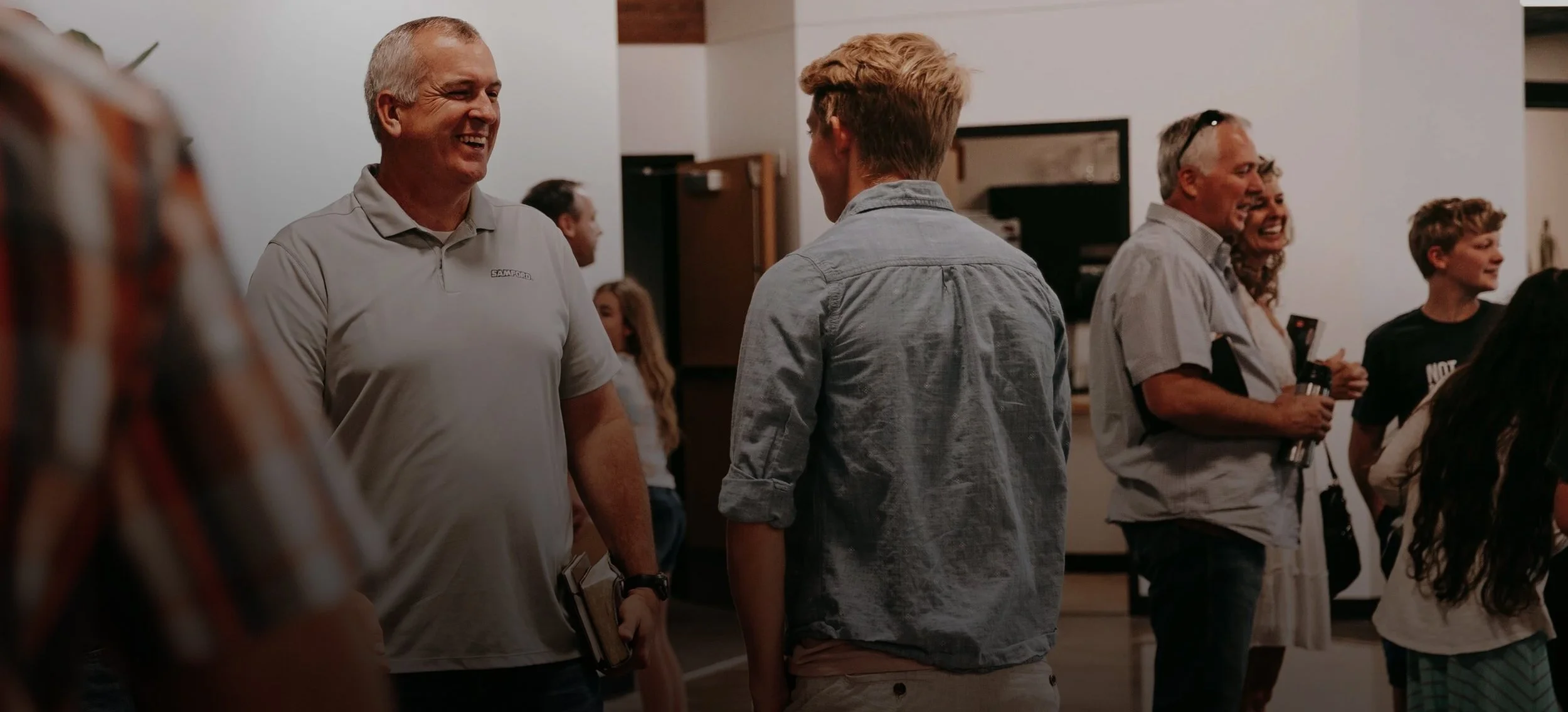 People smiling and talking in a social gathering indoors, with a man holding books and others holding drinks.