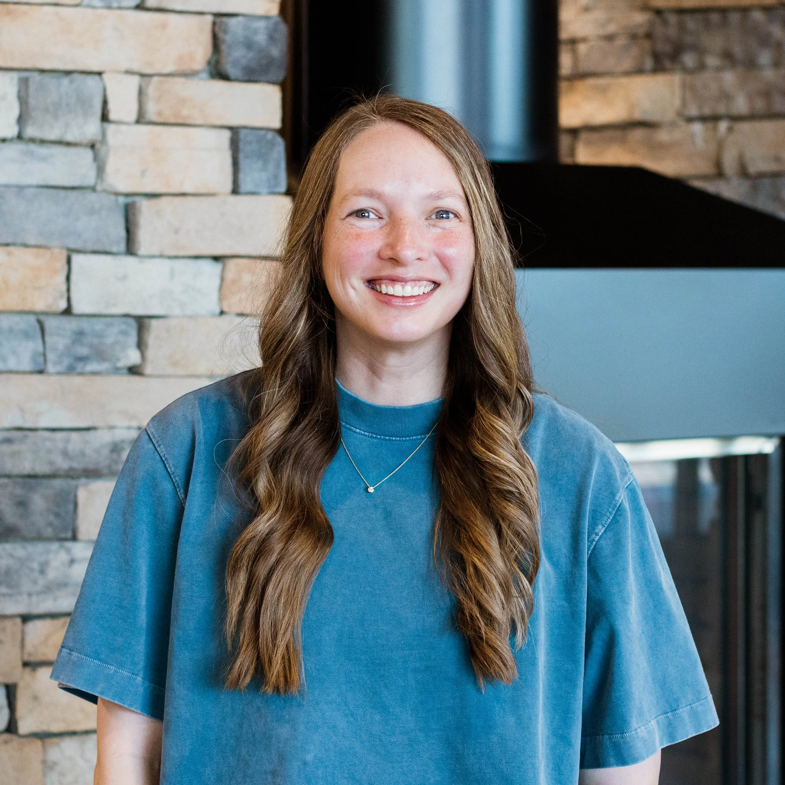 A young woman with long wavy hair smiling, wearing a black t-shirt, standing in front of a stone wall.