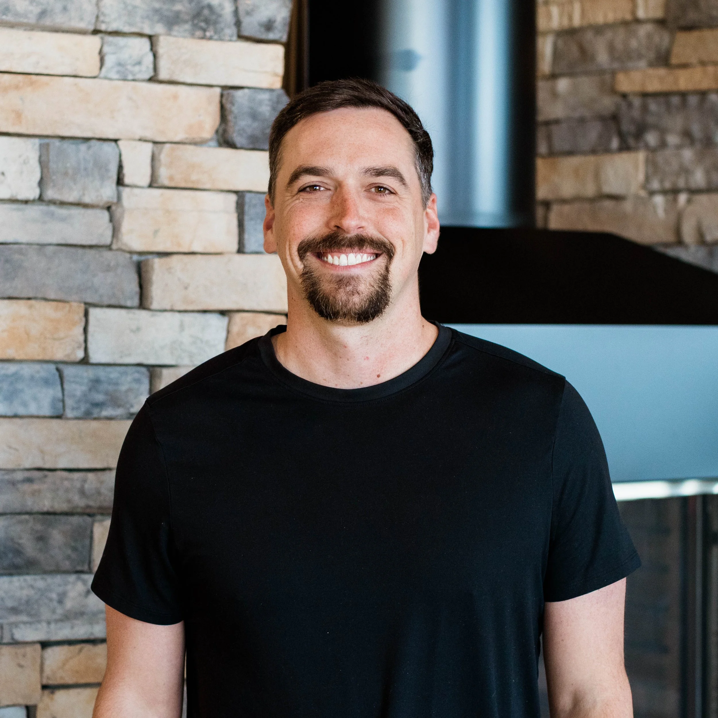 A man with dark hair and a goatee smiling, wearing a navy Under Armour hoodie, standing in front of a textured stone wall.