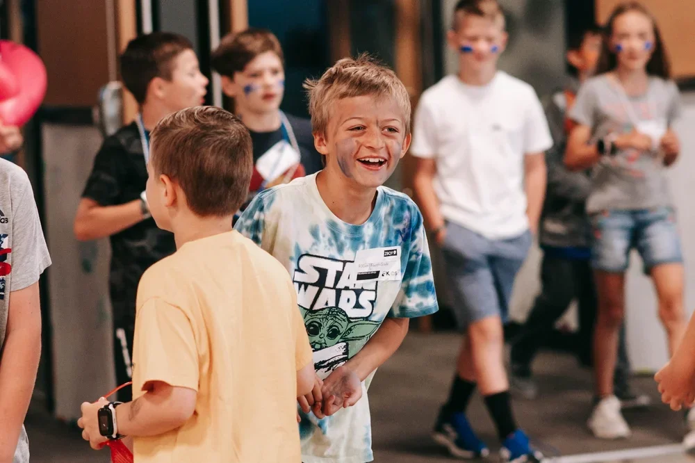 A group of children at a birthday party, with one boy in the foreground laughing and wearing a Star Wars T-shirt, surrounded by other kids with painted faces.