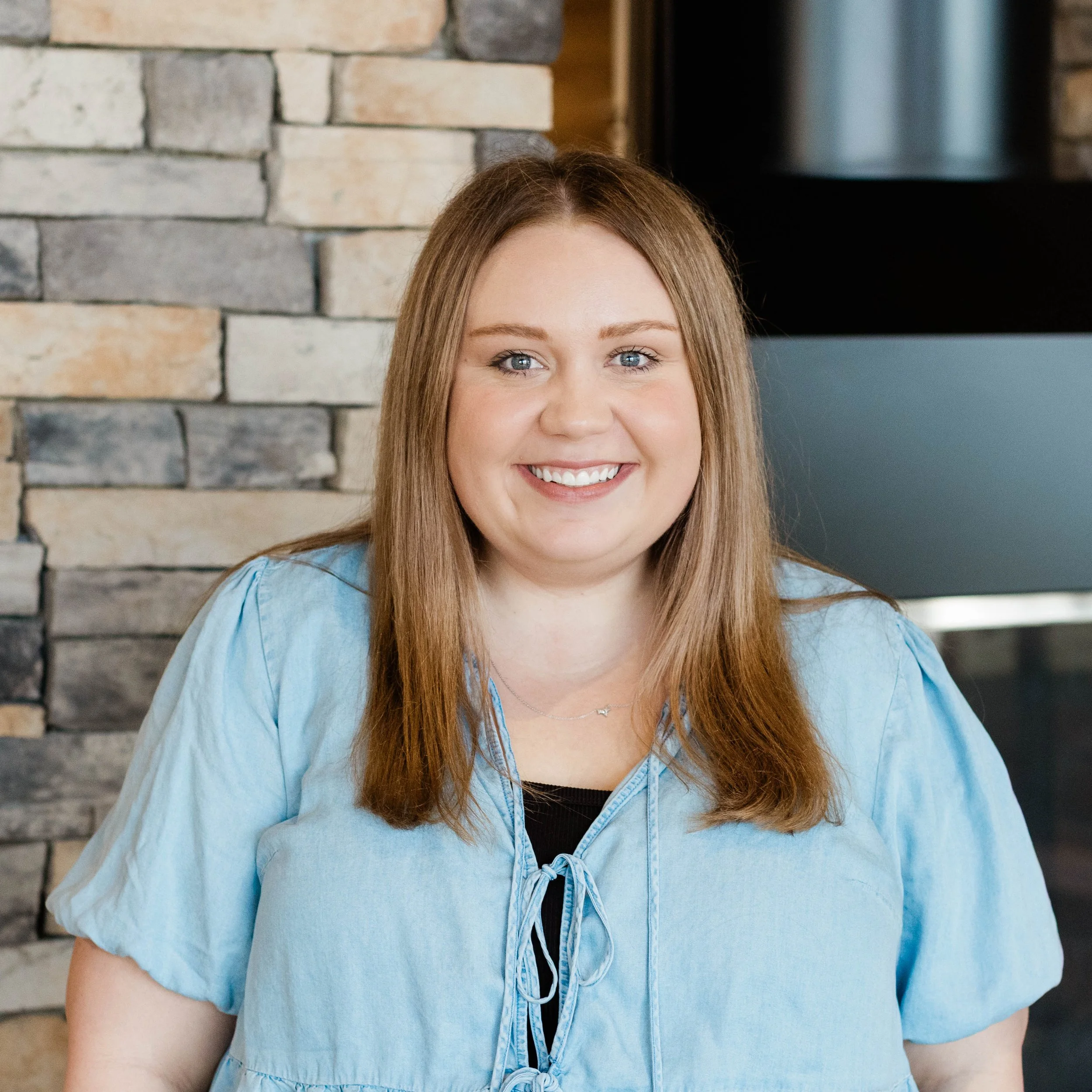 A smiling woman with wavy, shoulder-length blonde hair wearing a black dress with black tassel earrings, standing indoors next to a stone wall in a modern interior space.