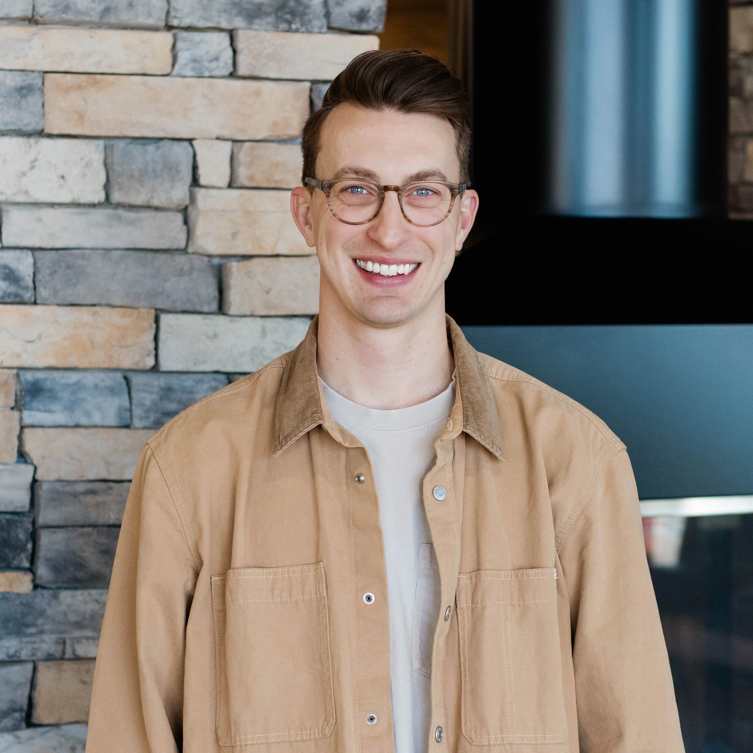 A smiling young man with glasses and short brown hair standing in front of a stone fireplace in a room with wooden furniture.