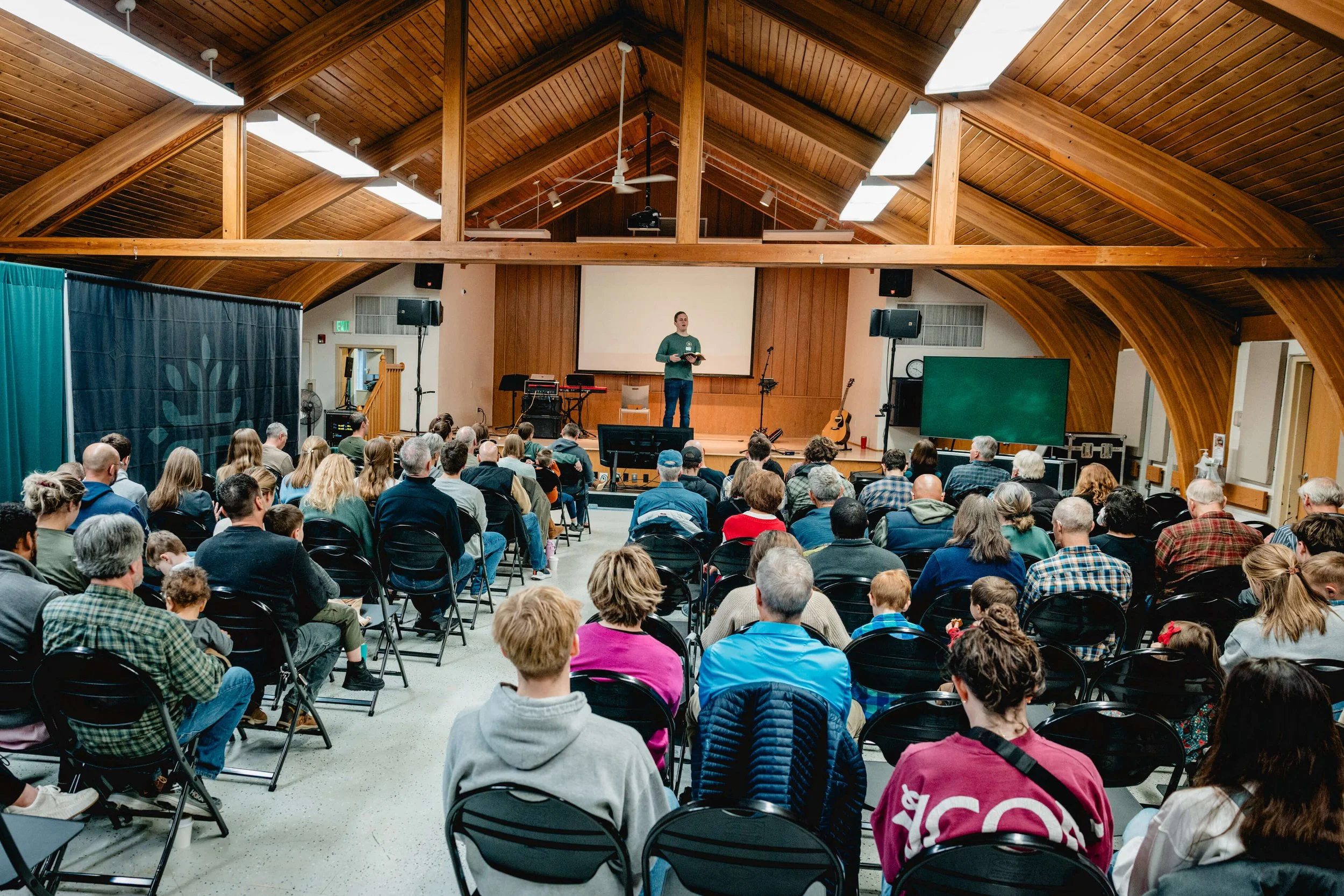 A large indoor gathering in a church or auditorium with a wooden arched ceiling, where a man on stage is addressing a seated audience. The stage has musical instruments, including a guitar and a keyboard, and a large screen behind him. Audience members are seated in black chairs, facing the stage, with some children and older adults present.