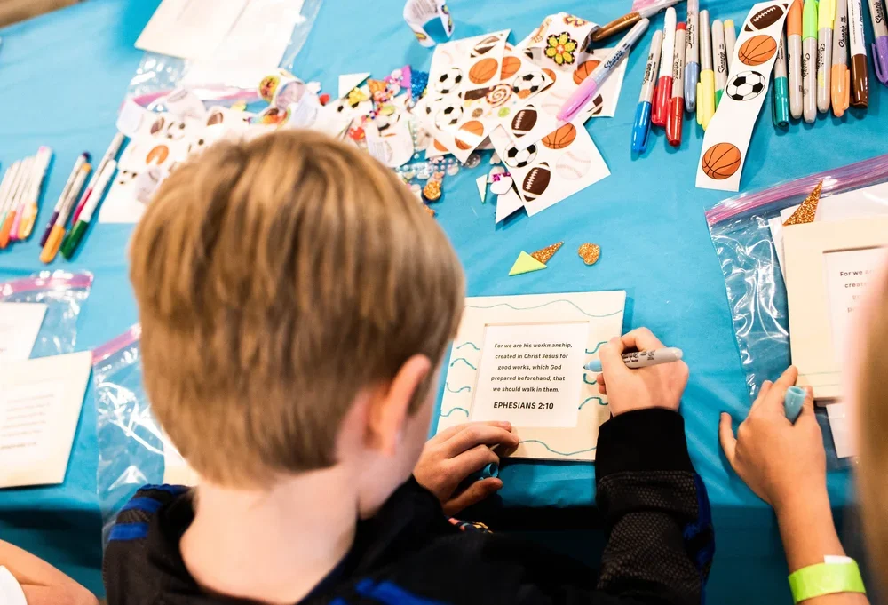 A child with light brown hair sitting at a table with craft supplies, coloring a religious card that displays a Bible verse, surrounded by colorful markers, stickers, and craft paper.