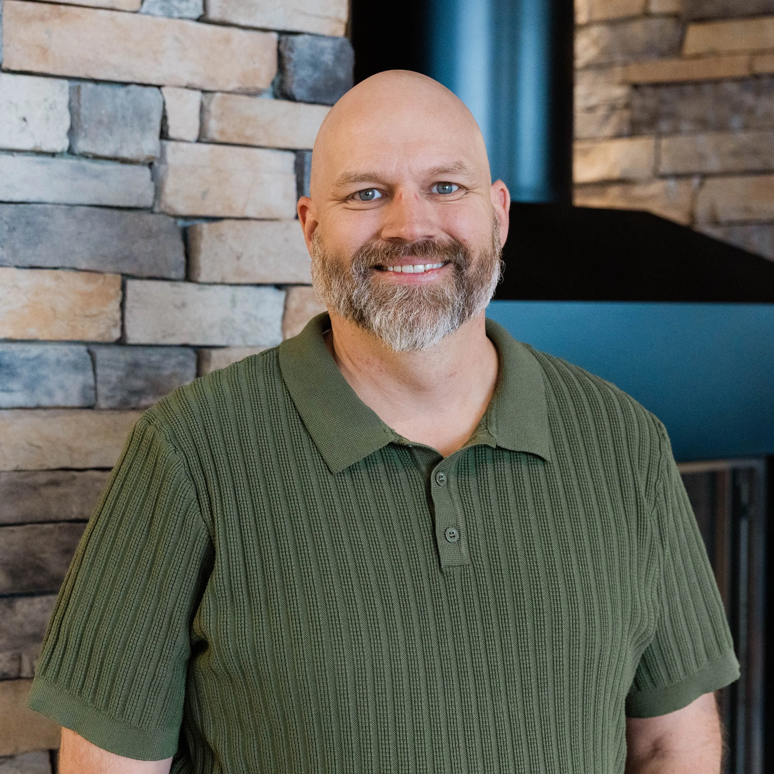 A smiling bald man with a beard and glasses, wearing a green shirt, standing in front of a stone fireplace.