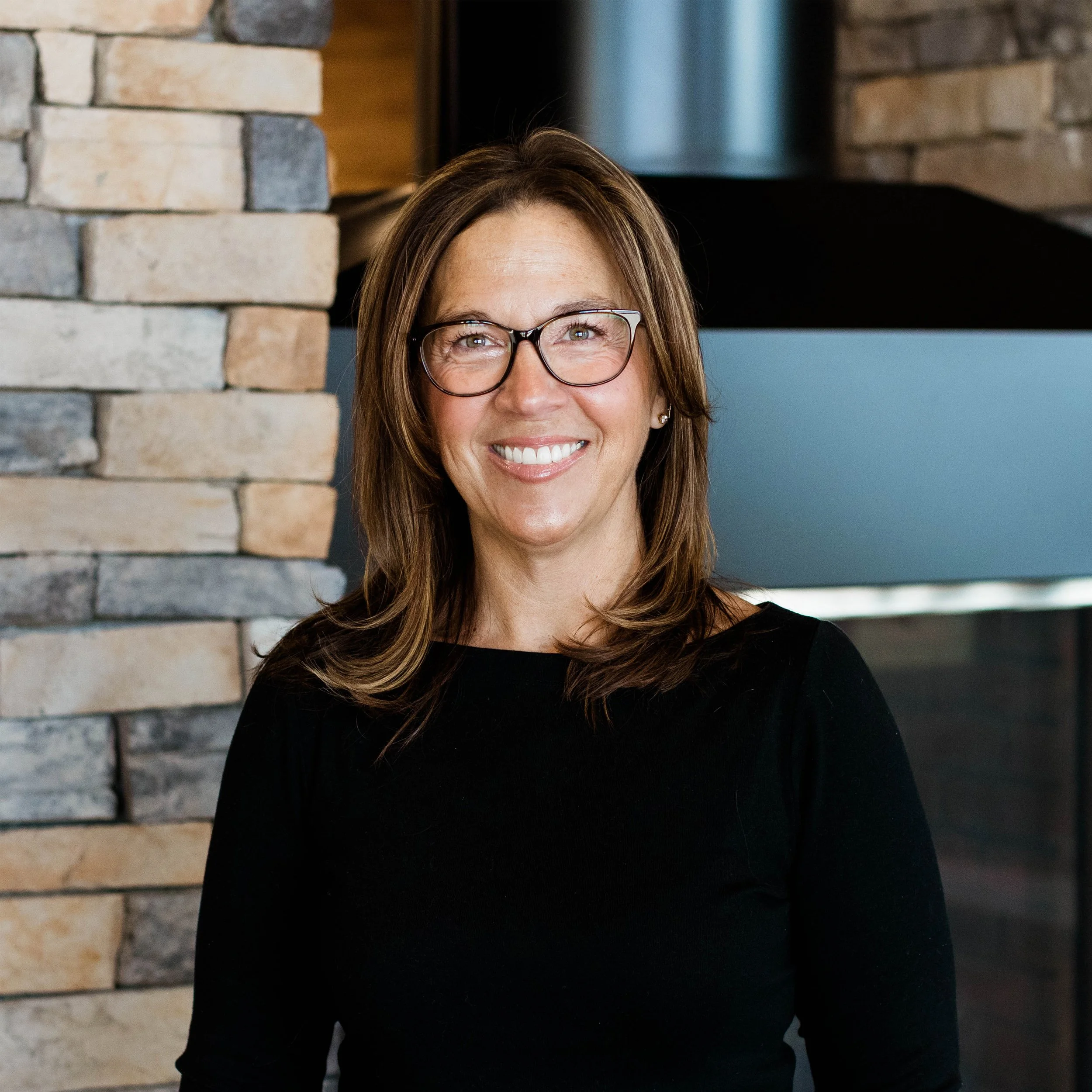 A woman with shoulder-length blonde hair, wearing glasses and a black shirt, smiling in front of a stone fireplace.