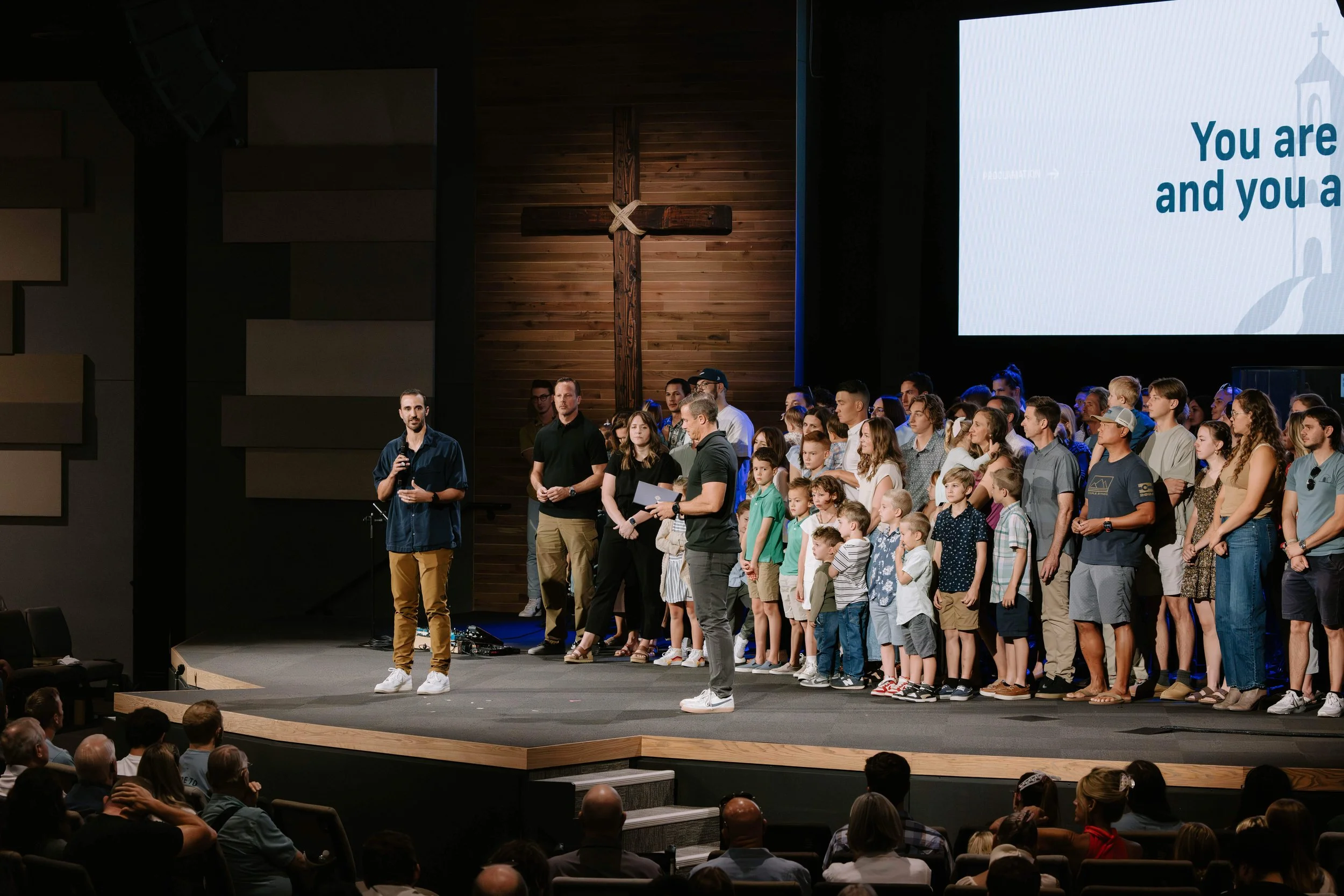 A large group of people, including children and adults, standing on a stage inside a church, with a large wooden cross on the wall behind them. A man in a blue shirt is speaking into a microphone, and others are gathered around him. Audience members are seated in front. A large screen displays partial text: 'You are and you a'.