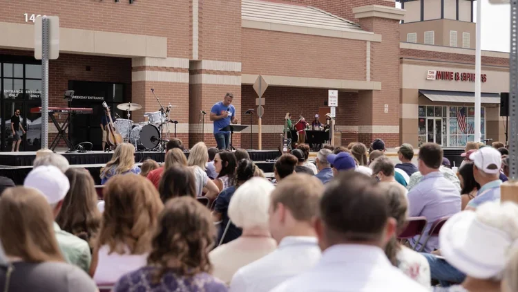 An outdoor stage with a man speaking into a microphone in front of an audience at a shopping center. The stage has musical instruments, and there are various people and storefronts in the background.