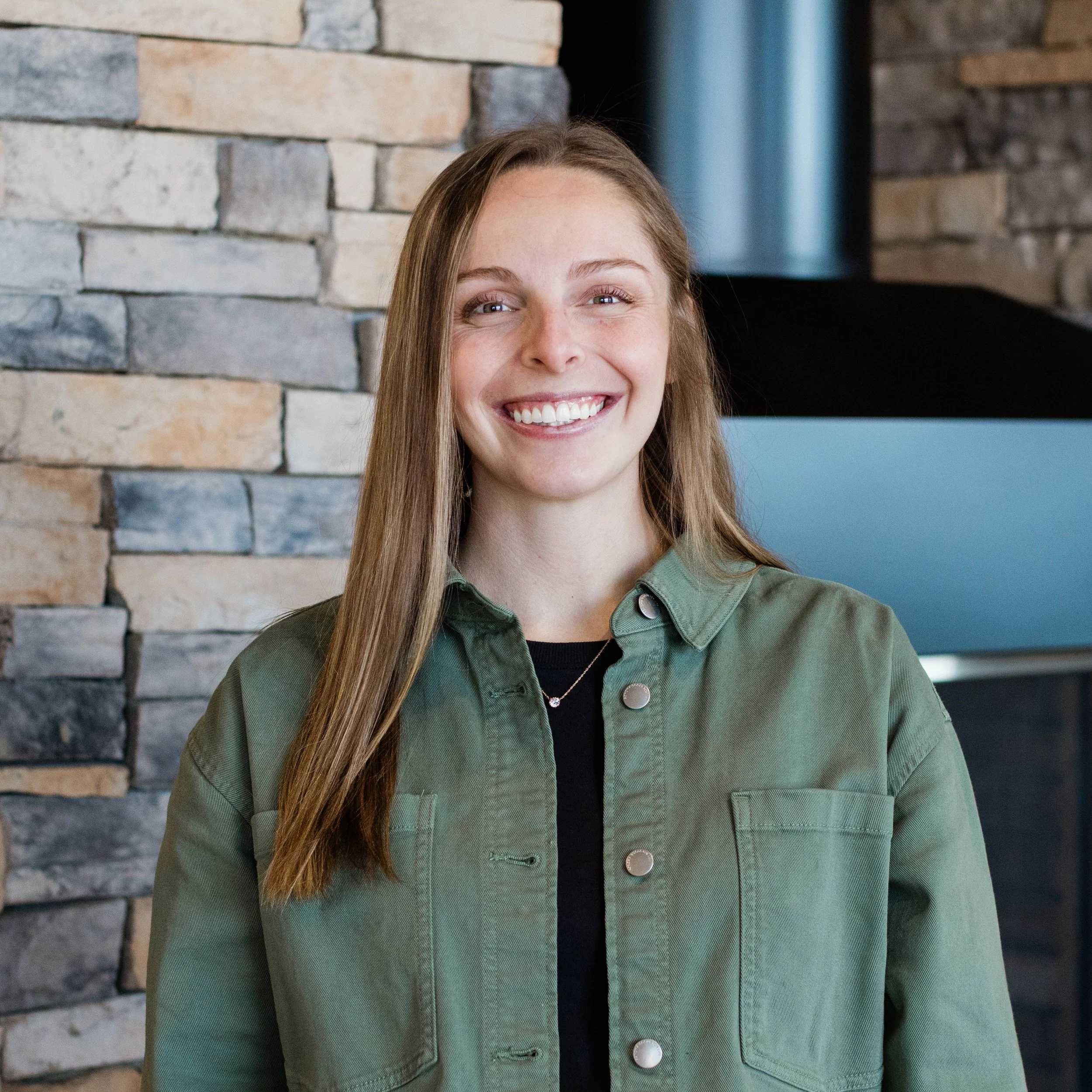 A young woman with long brown hair, smiling, wearing an orange blouse, standing indoors near a stone wall.