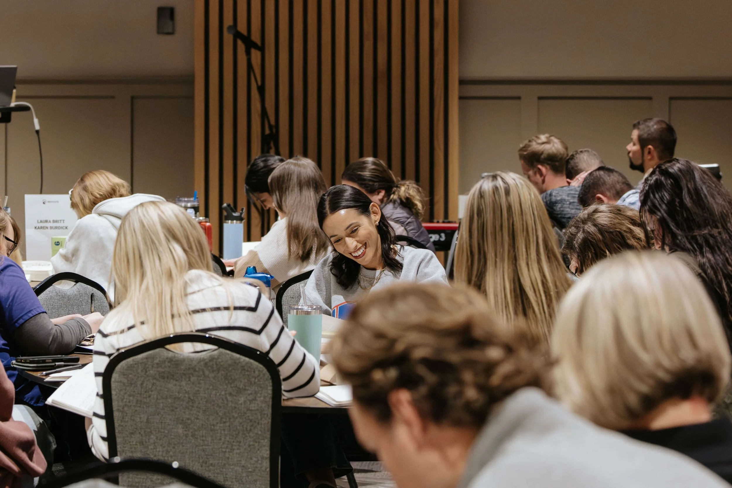 A group of people seated at round tables in a conference room, engaging and smiling during a workshop or seminar.