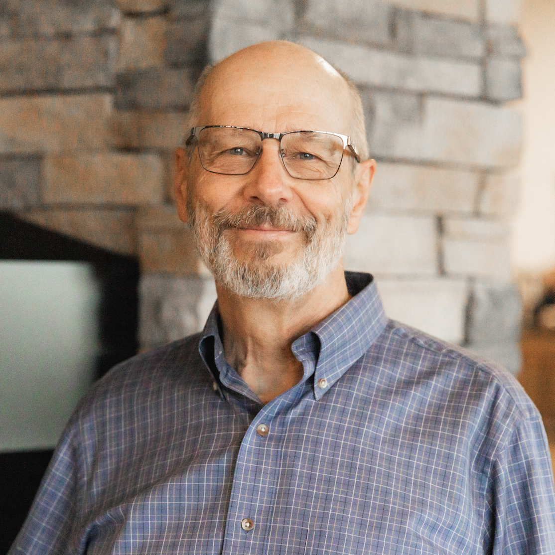 A smiling elderly man with a beard and glasses, wearing a plaid button-up shirt, standing in front of a stone fireplace.