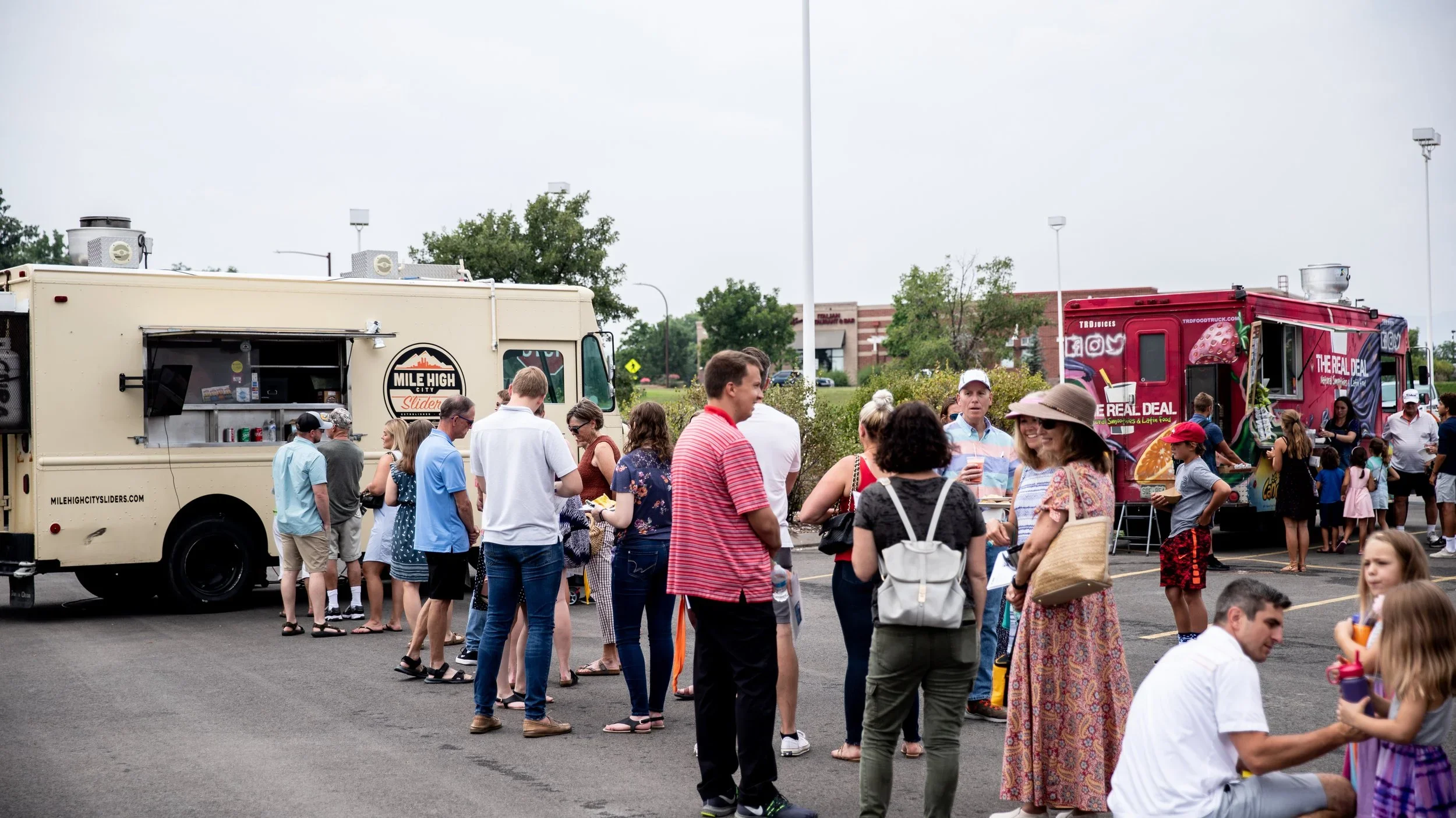 People standing in line outside two food trucks in a parking lot on a cloudy day, with a building and trees in the background.