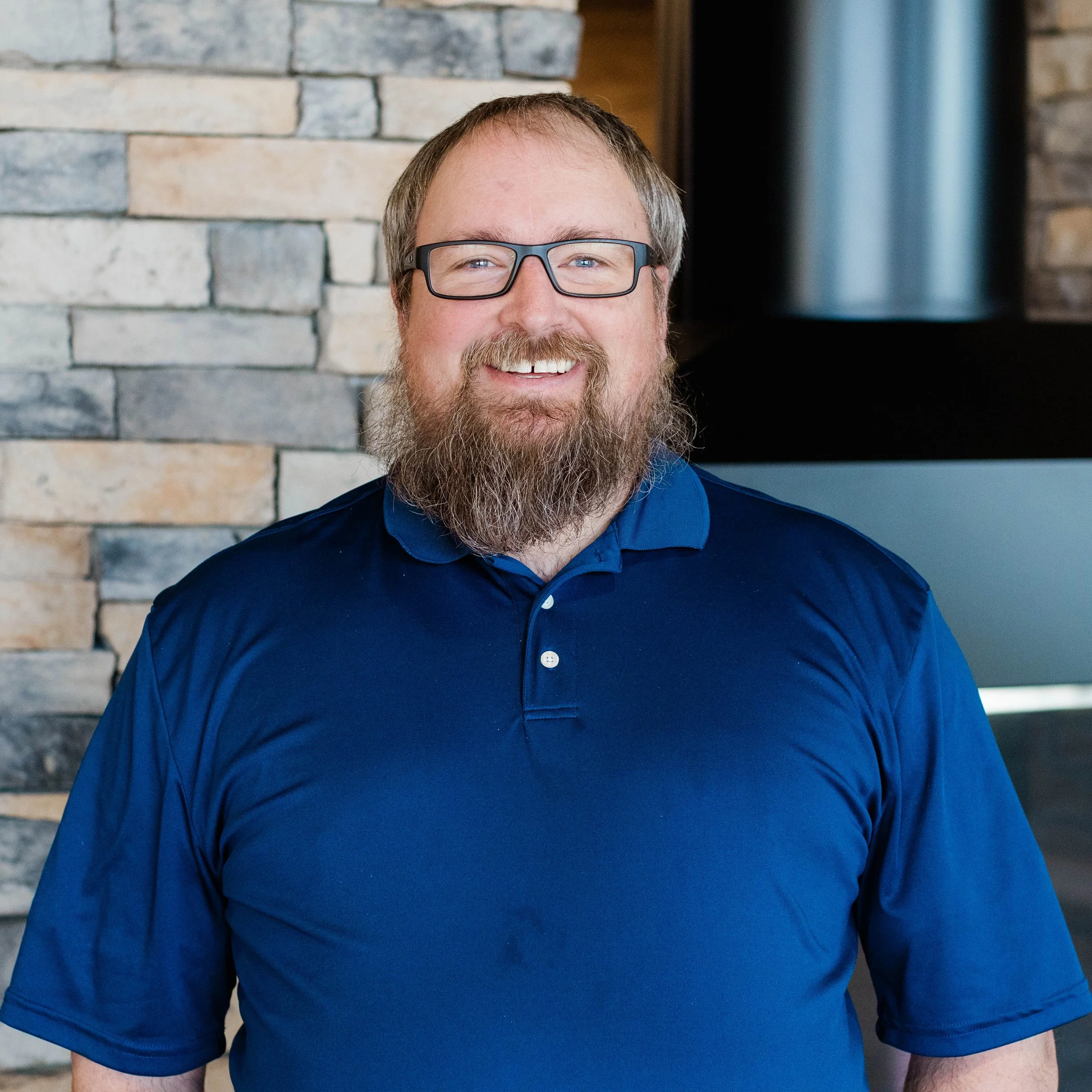 A smiling man with a beard wearing a black polo shirt standing indoors near a stone wall.