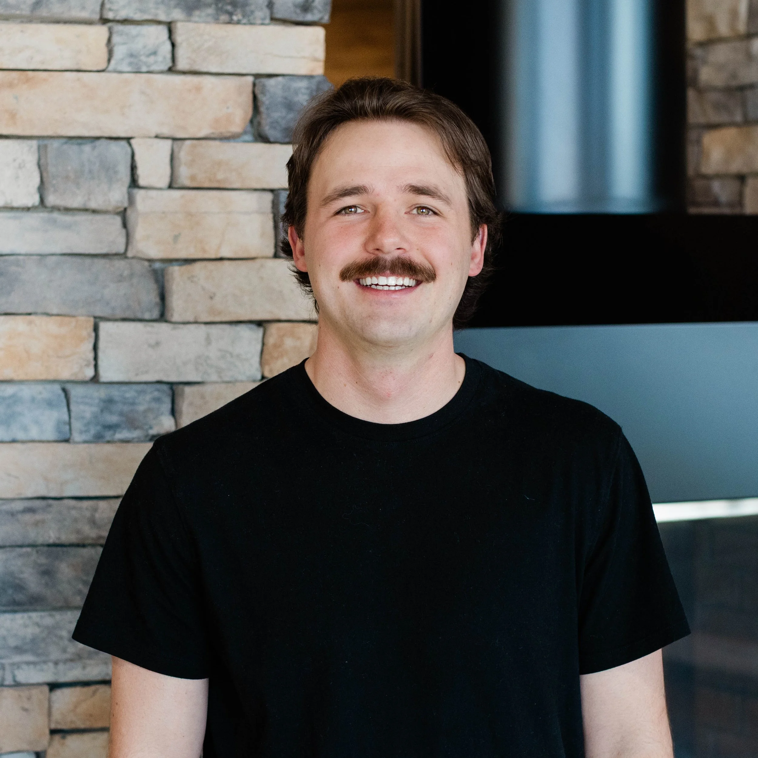 Smiling man with a mustache and dark hair, wearing a black t-shirt, standing in front of a stone wall near a fireplace.