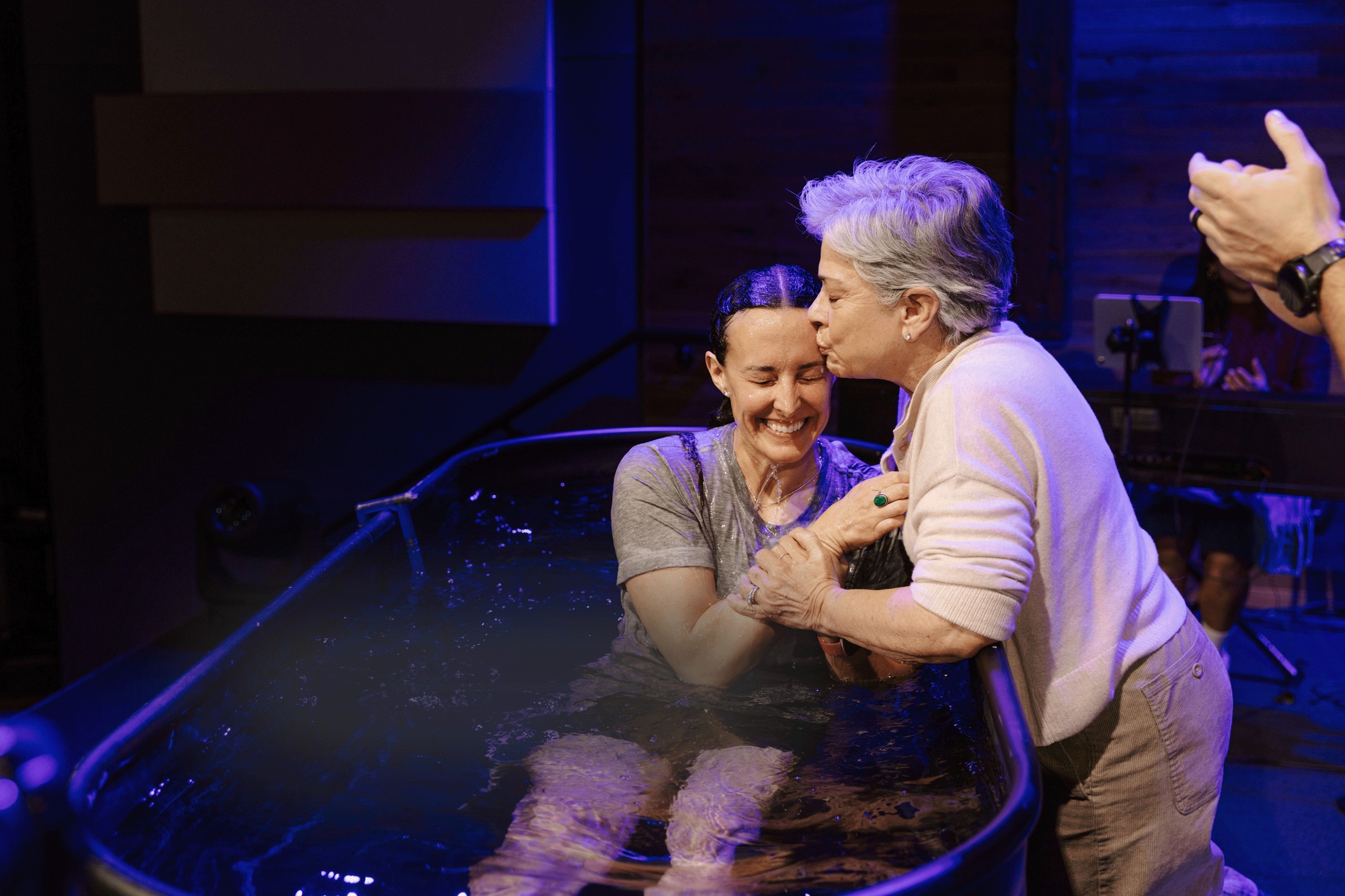 A group of people at a Christian baptism, with three men in the foreground inside a baptismal pool, one being baptized and two others praying and celebrating.