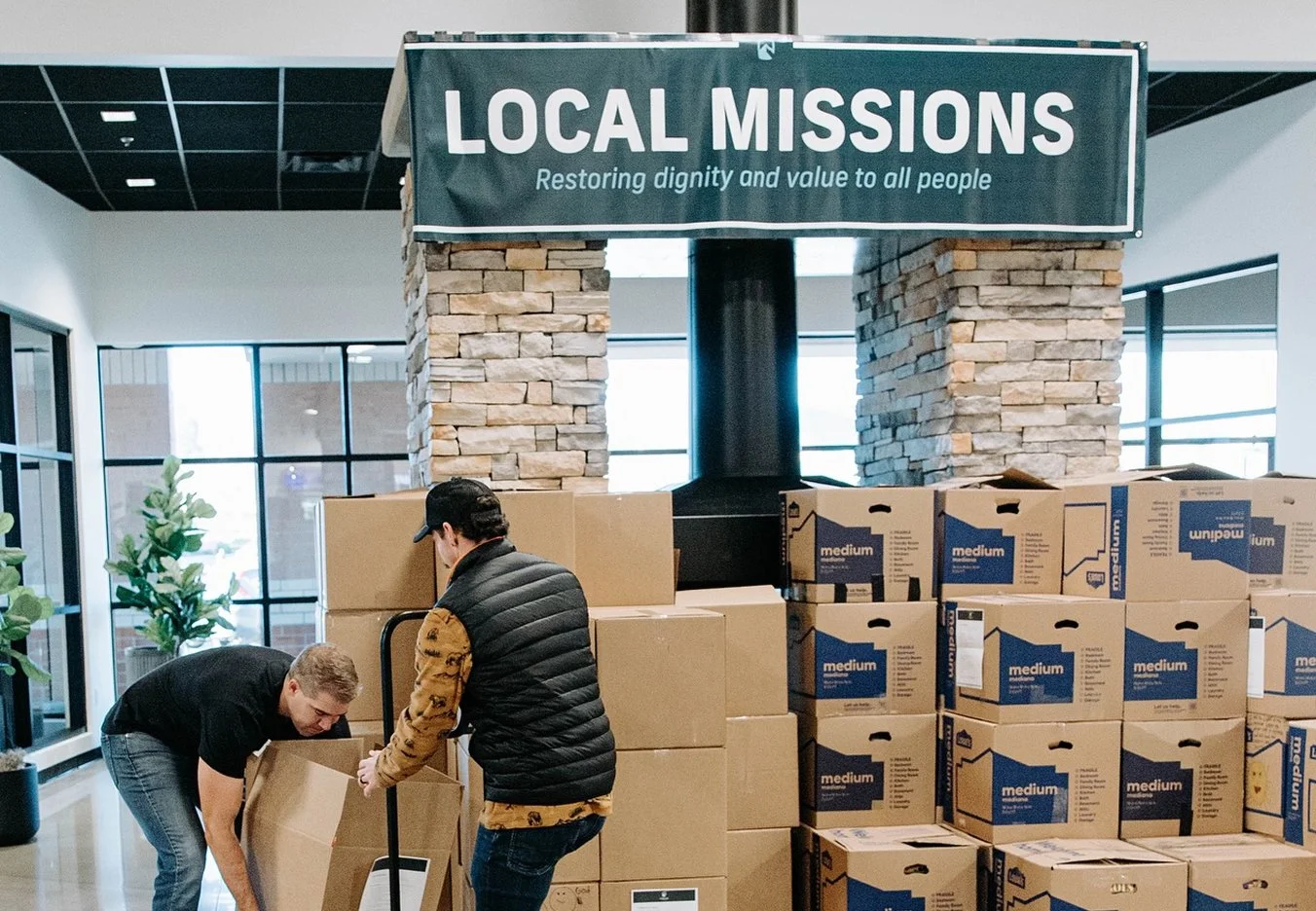 Two men unload boxes of supplies in a warehouse or donation center, with a sign above reading 'Local Missions: Restoring dignity and value to all people.'