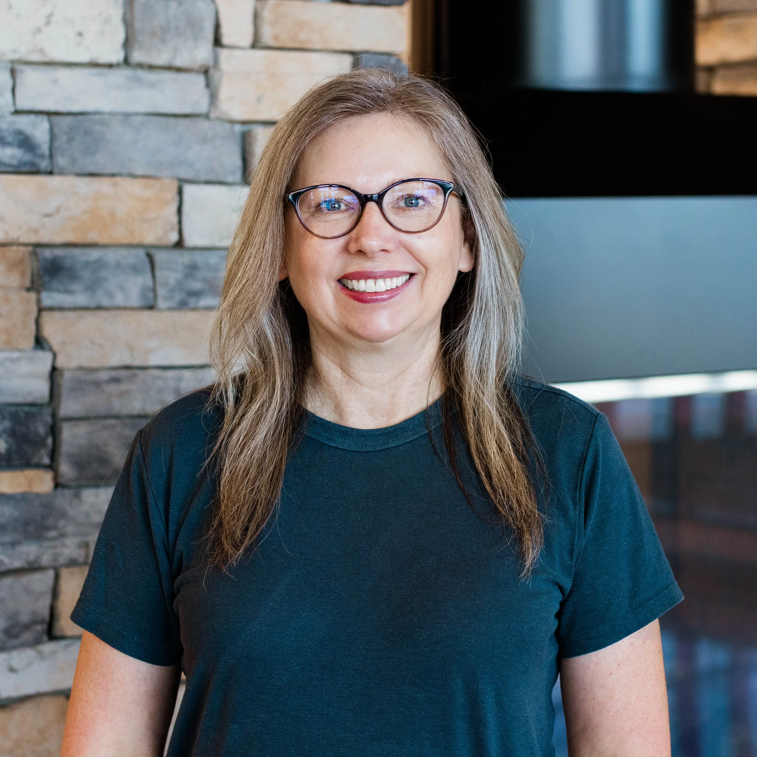 A woman with long brown hair, glasses, and a slight smile standing in front of a stone wall with a fireplace.