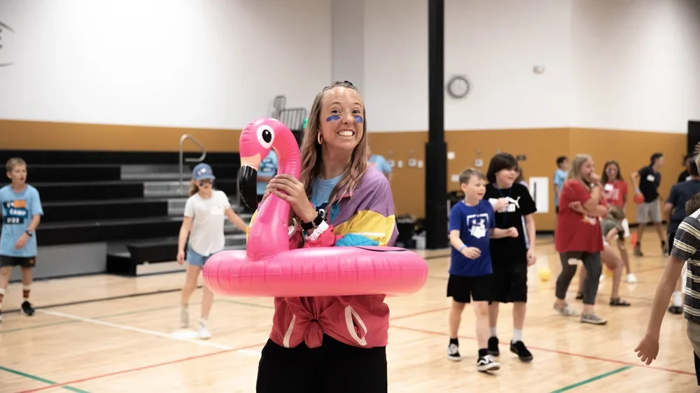A woman wearing a colorful jacket and face paint is holding a pink inflatable flamingo in a gymnasium filled with children.