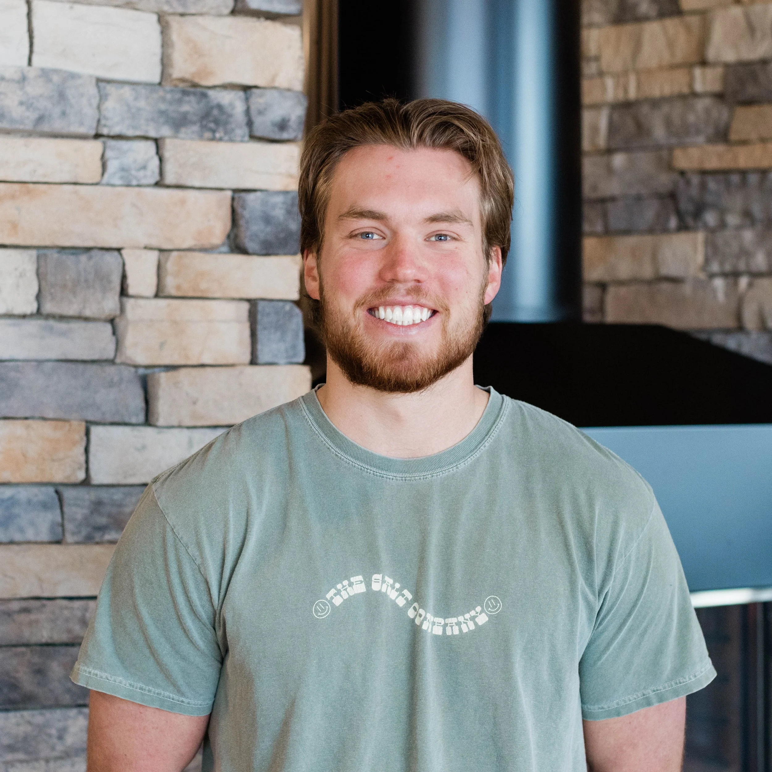 A young man with brown hair and a beard smiling, wearing a light-colored t-shirt, standing in front of a stone fireplace with stacked stone pattern.