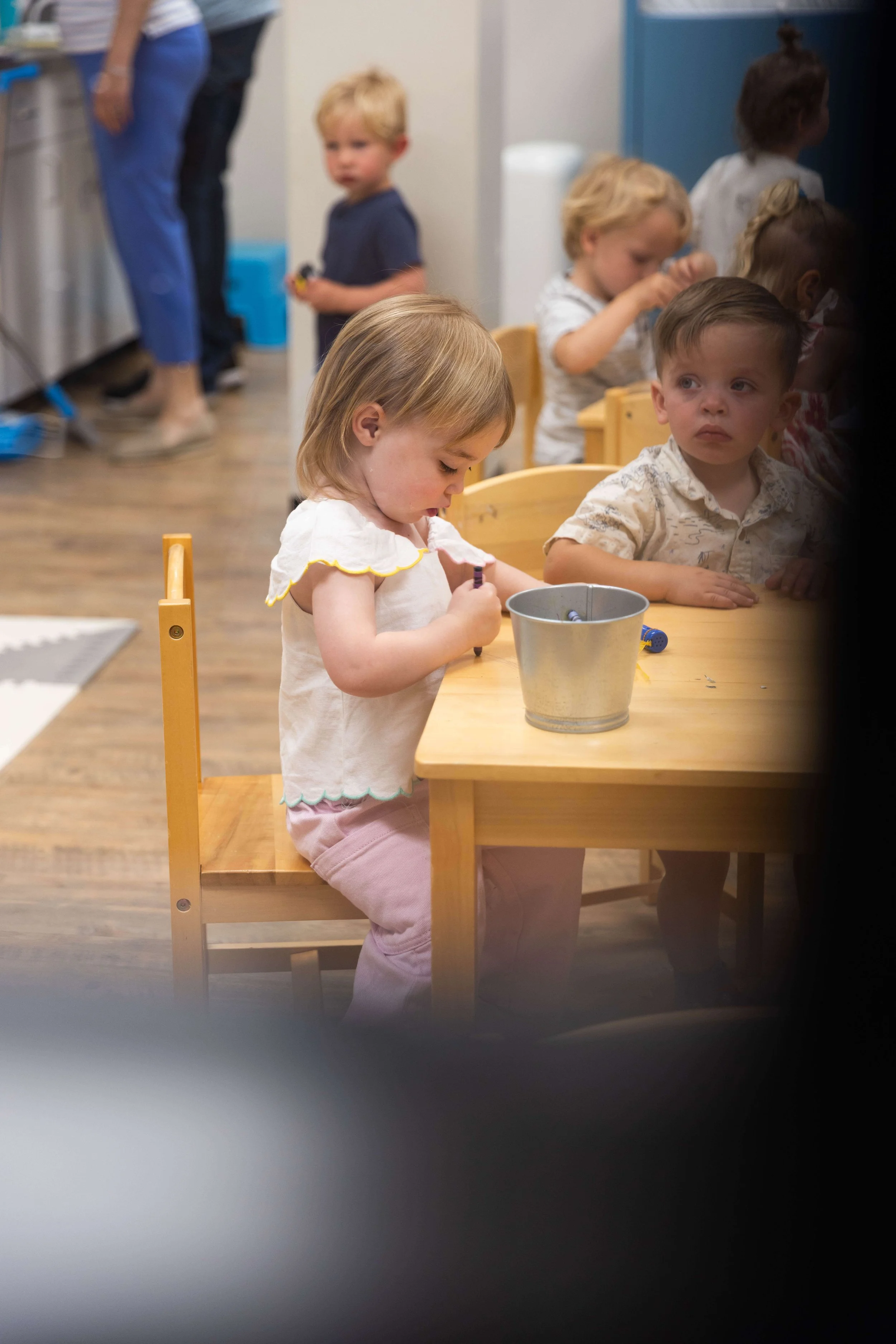 Children sitting and standing around a wooden table, engaging in various activities in a classroom or daycare setting.