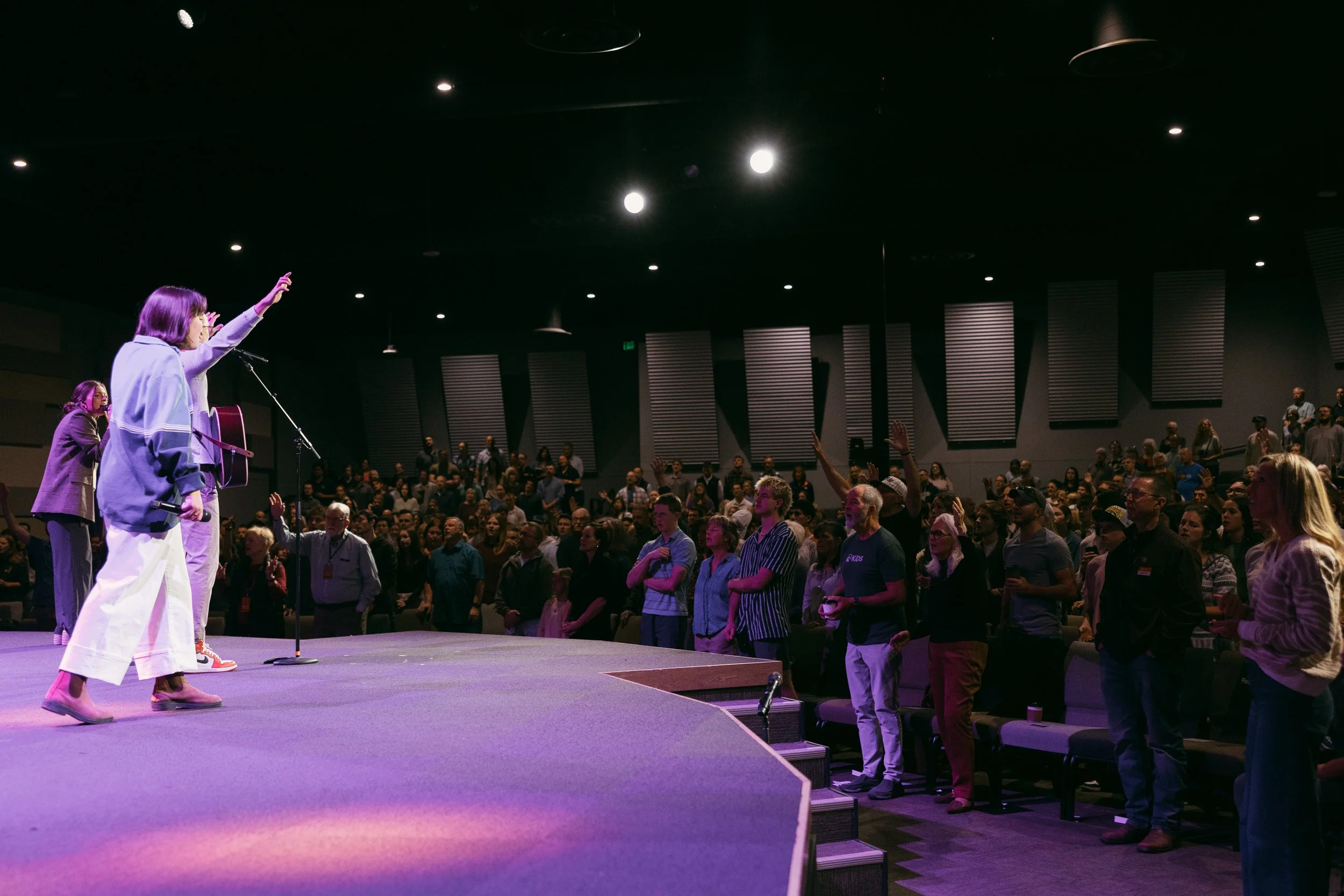 People standing and raising their hands during a church service or event, with two women on stage leading the congregation.