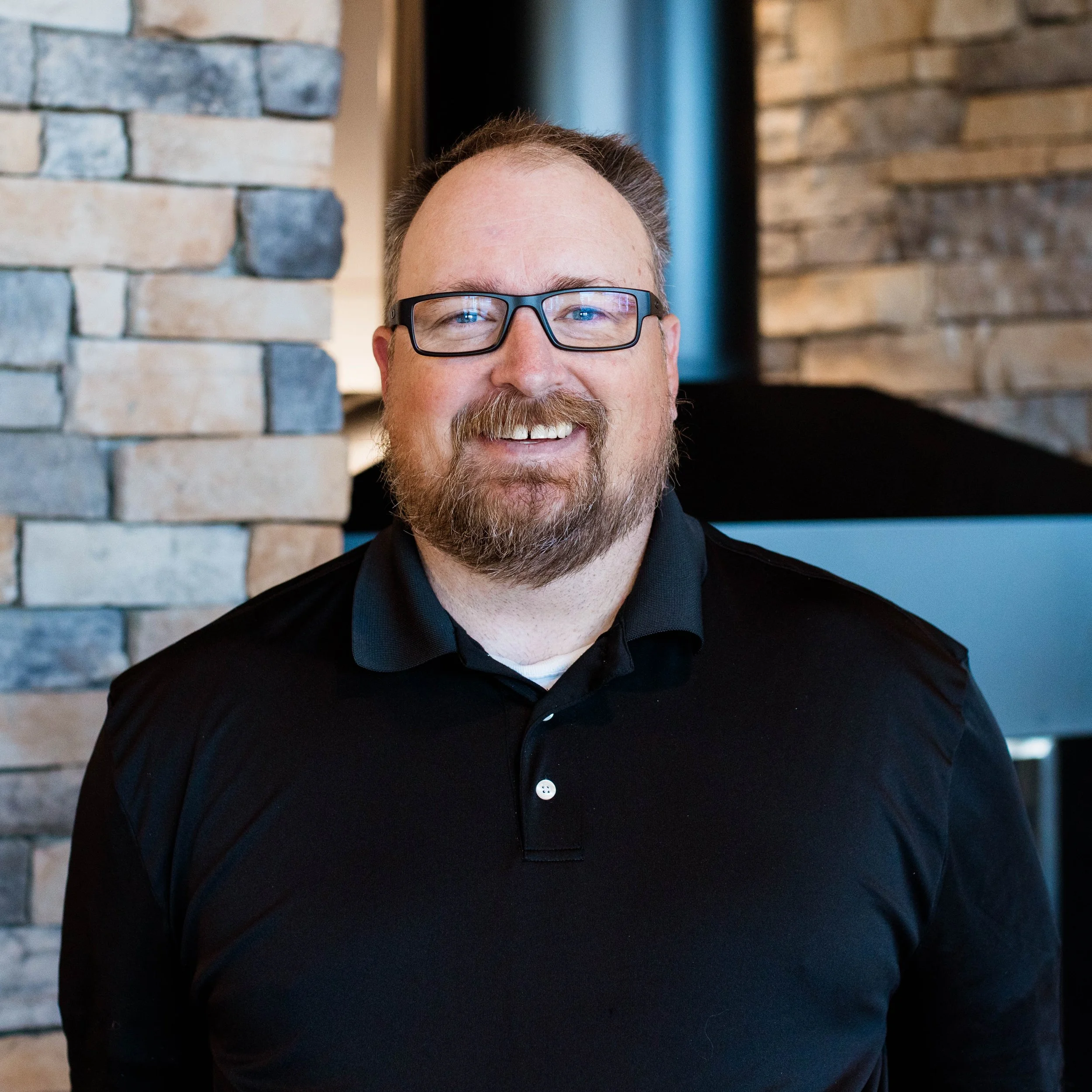 A smiling man with a beard wearing a black polo shirt standing indoors near a stone wall.