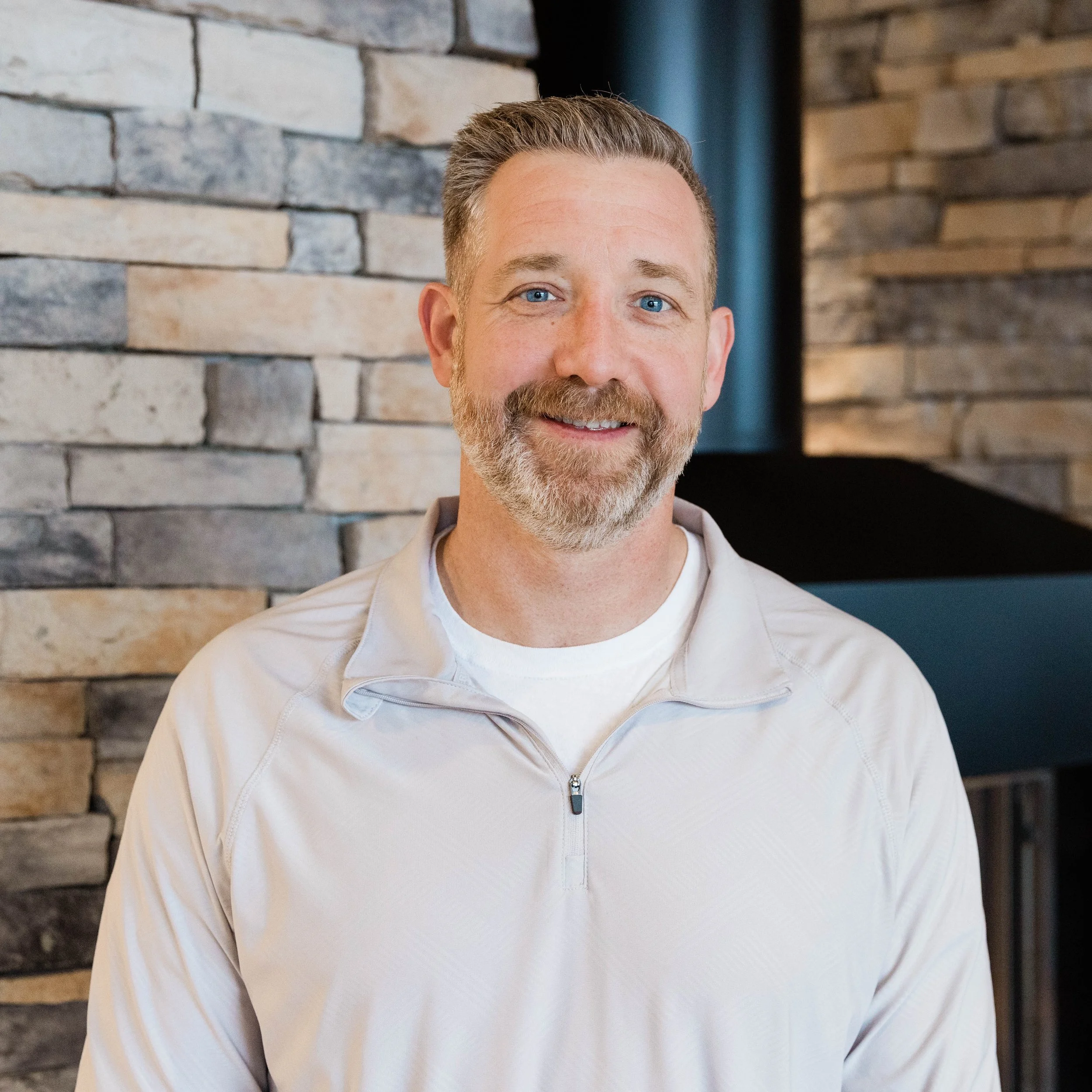 A smiling middle-aged man with a beard, wearing a gray jacket over a white shirt, standing in front of a brick wall near a fireplace.