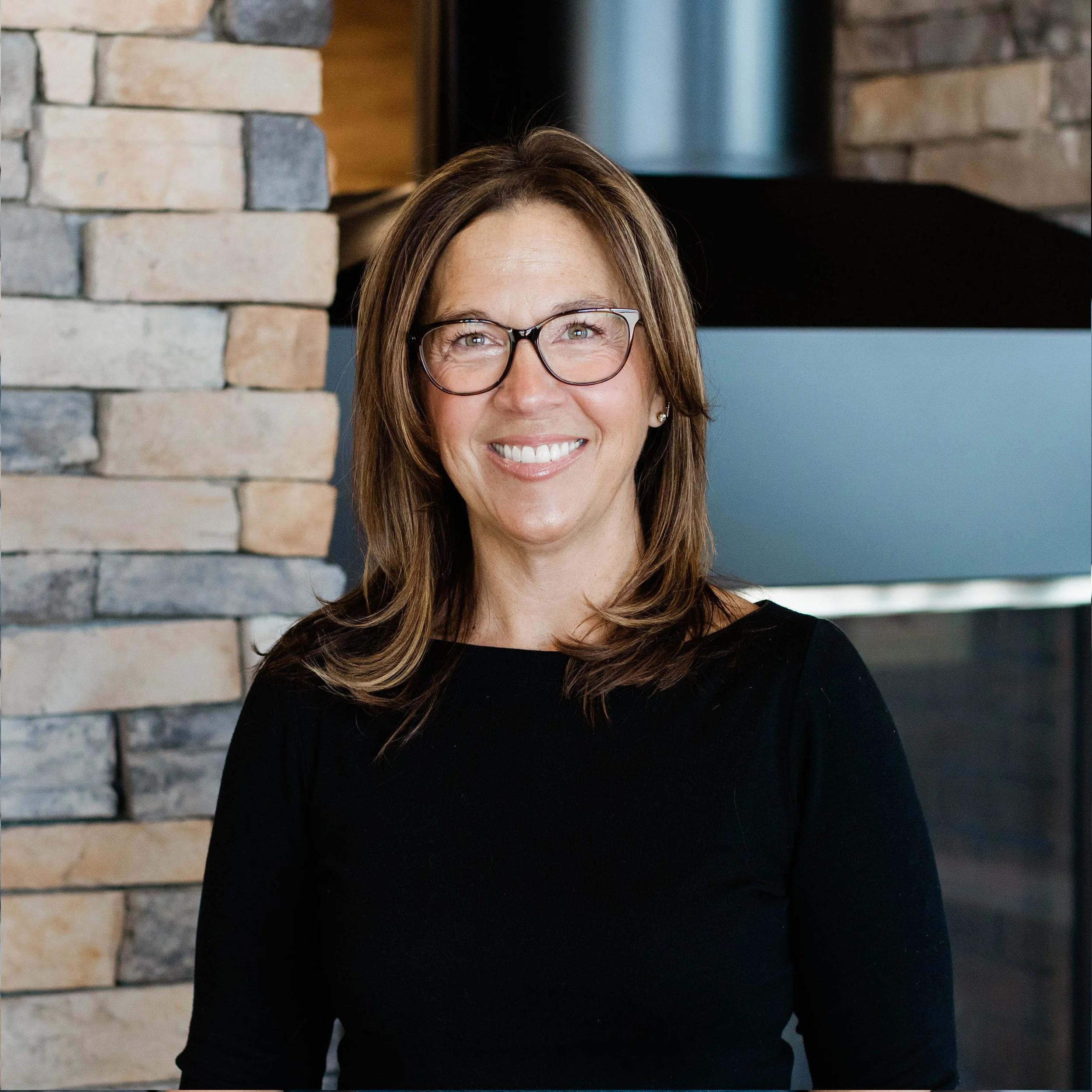 A woman with shoulder-length blonde hair, wearing glasses and a black shirt, smiling in front of a stone fireplace.