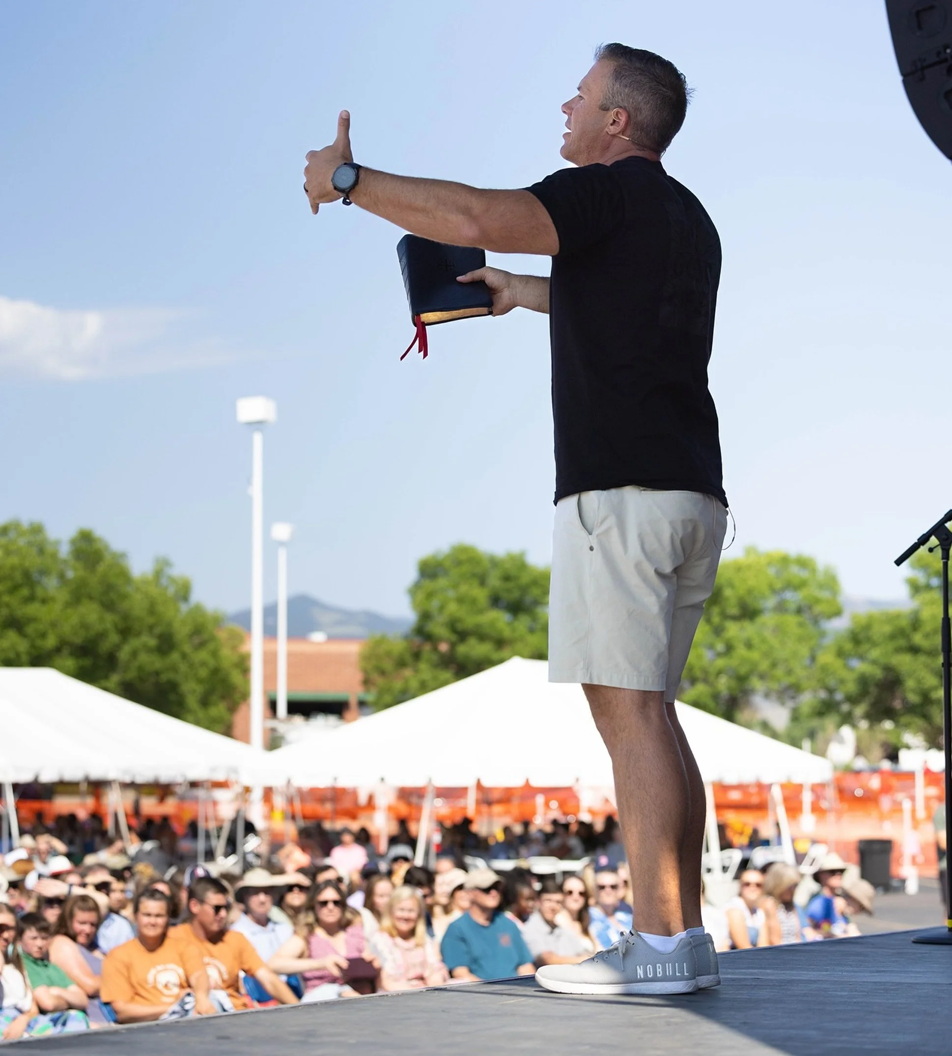 A man in a black t-shirt, beige shorts, and white sneakers is standing on an outdoor stage, holding a book in one hand and gesturing with the other during a speech or presentation. Behind him is an audience seated in front of tents, with trees and a clear sky in the background.