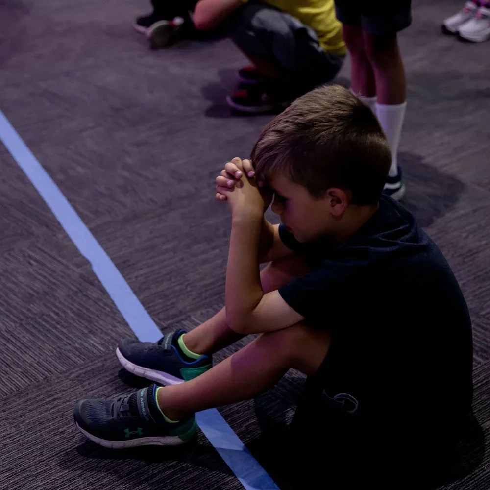 A young boy sitting on the floor with his head bowed and his hands clasped in front of his forehead, appearing to be in prayer or deep thought, in an indoor setting with other children around.
