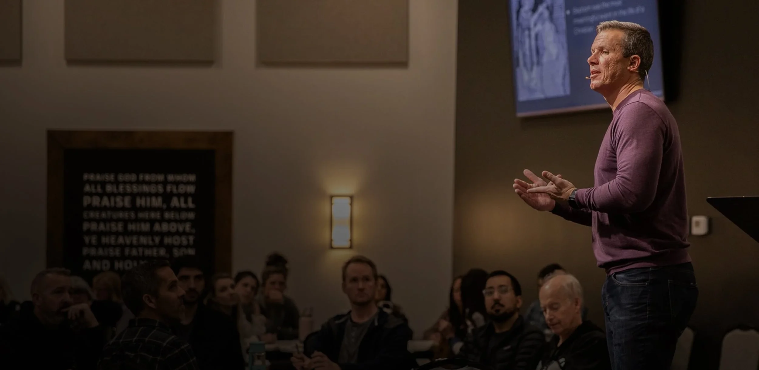 A man giving a talk or presentation in front of an audience in a dimly lit room. The man is wearing a purple long-sleeve shirt and has a microphone headset. The audience is seated and attentive, with a large screen displaying information behind the speaker, and a framed text on the wall that says praise to God.