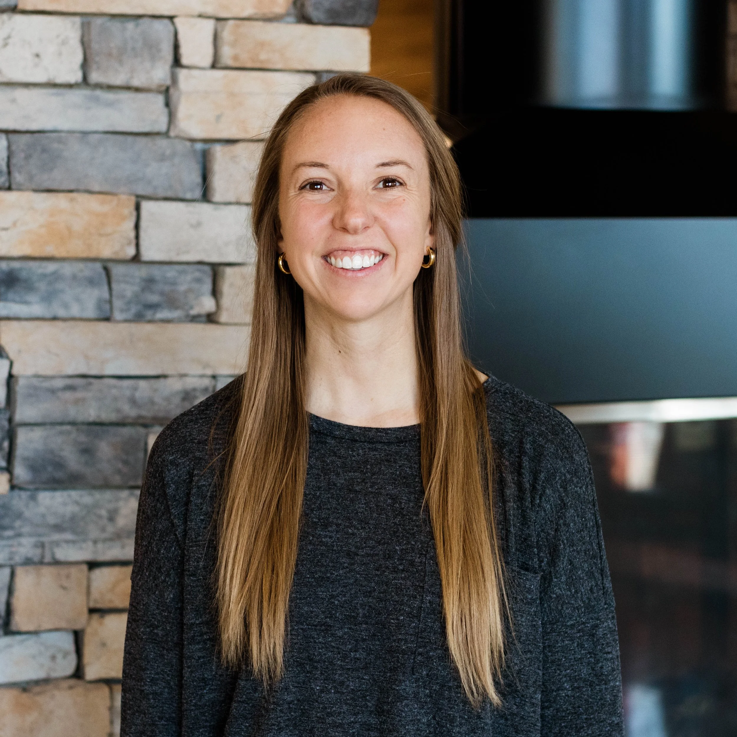 Smiling woman with long blonde hair standing in front of a stone fireplace wall.