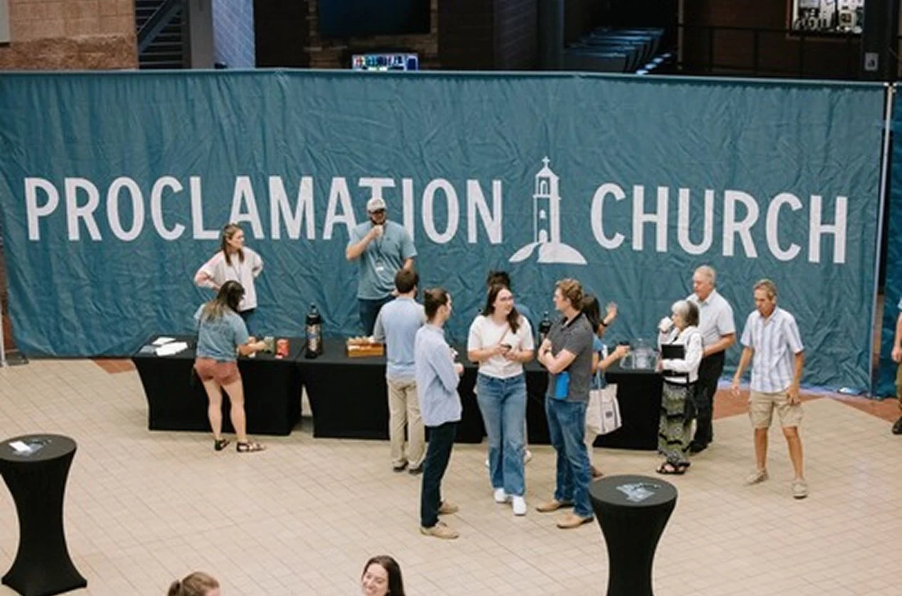 People gathered for a church proclamation event in an indoor venue with a large blue banner that says 'Proclamation at Church' and features a church tower illustration. Some are standing in line at refreshment tables, while others are engaged in conversation.