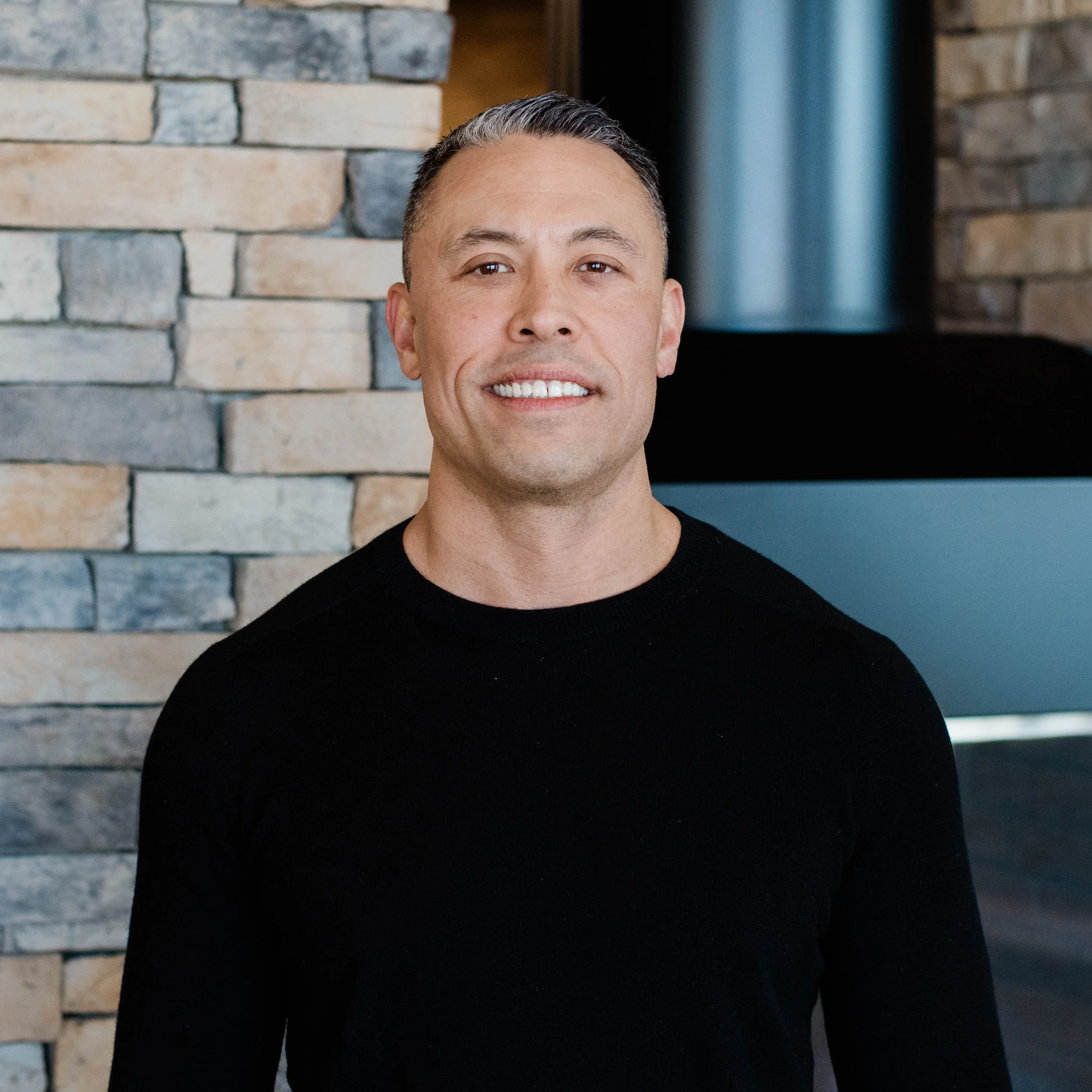 A smiling man with short dark hair and a goatee standing indoors in front of a stone wall and fireplace.