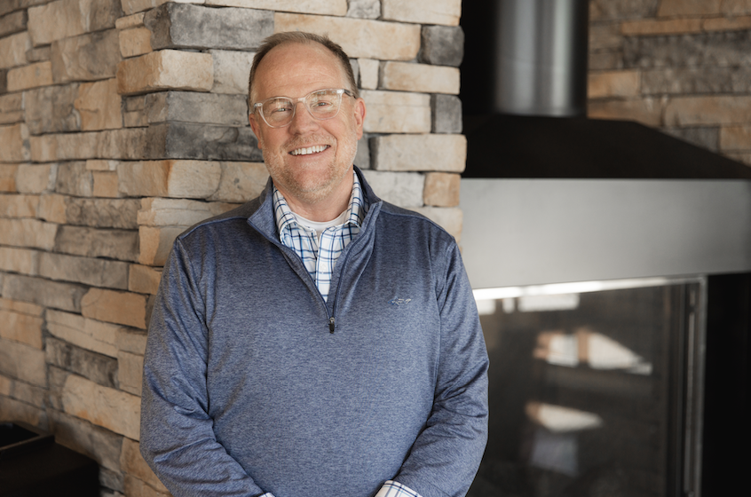 A smiling middle-aged man with glasses and a beard, wearing a blue jacket over a plaid shirt, standing in front of a brick wall near a fireplace.