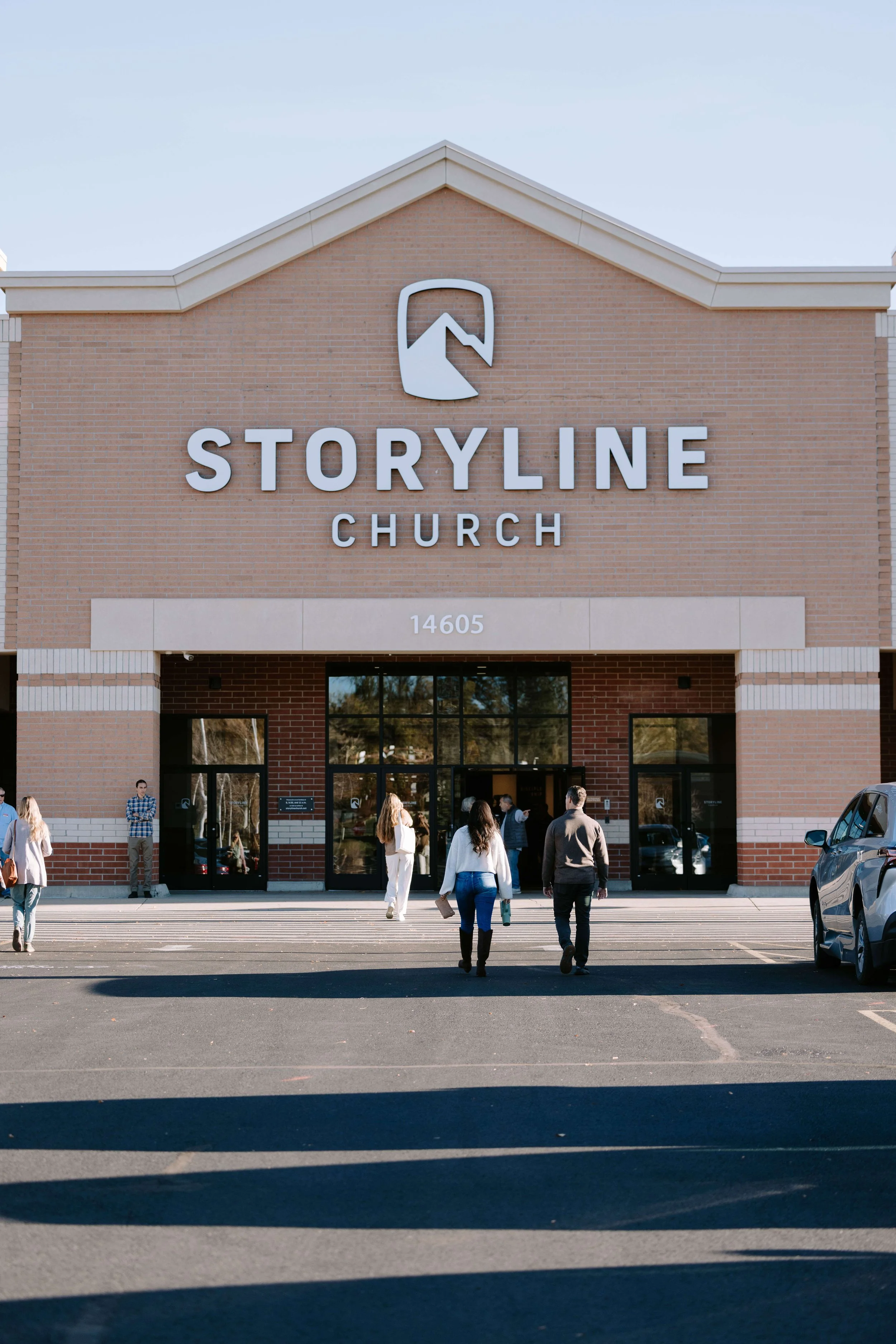 Front view of Storyline Church building with people walking towards entrance, parking lot in foreground, and clear sky above.