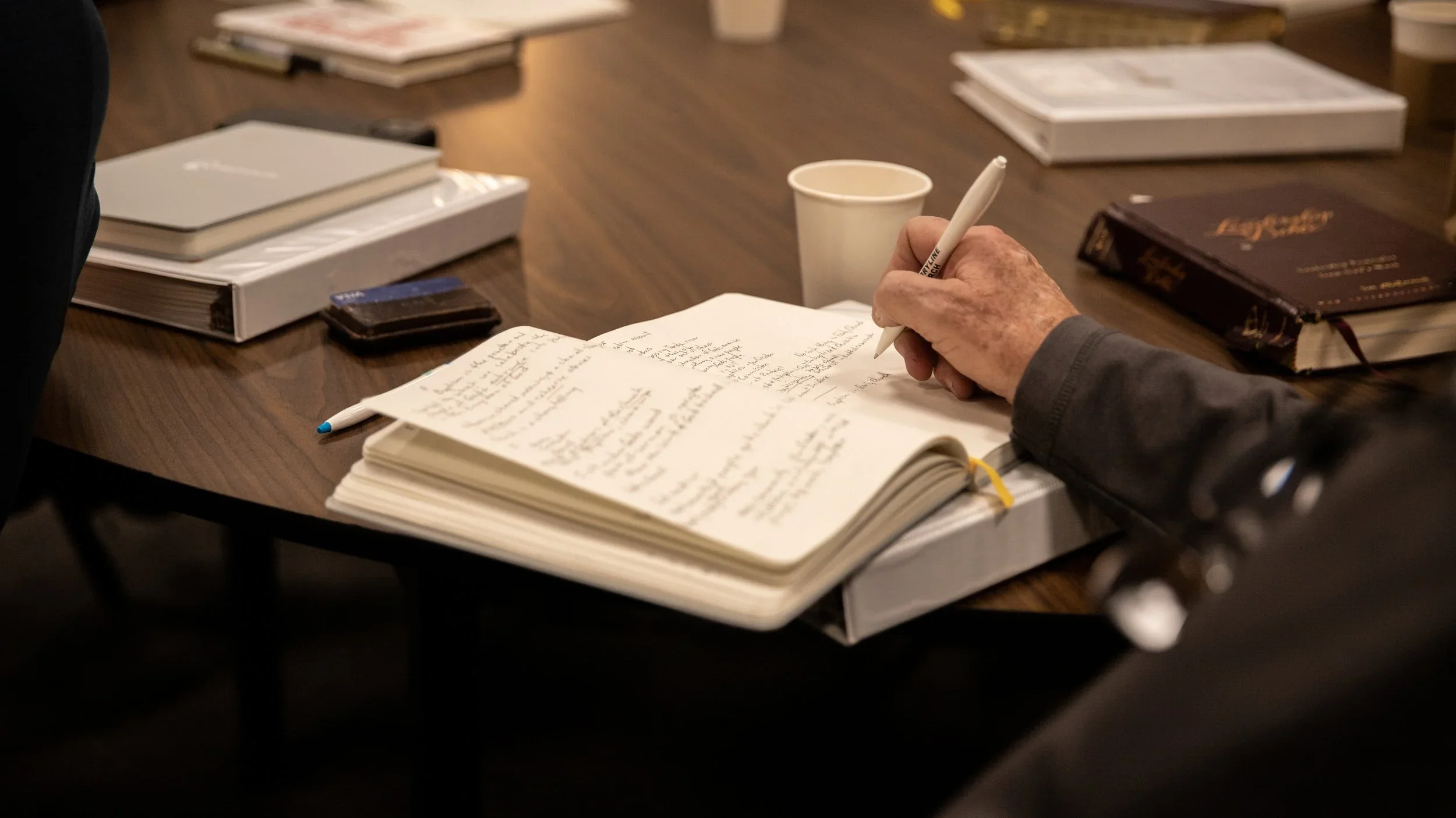 Close-up of an elderly person's hand holding a pen and writing in a notebook on a wooden table, surrounded by books, a coffee cup, and a closed laptop.