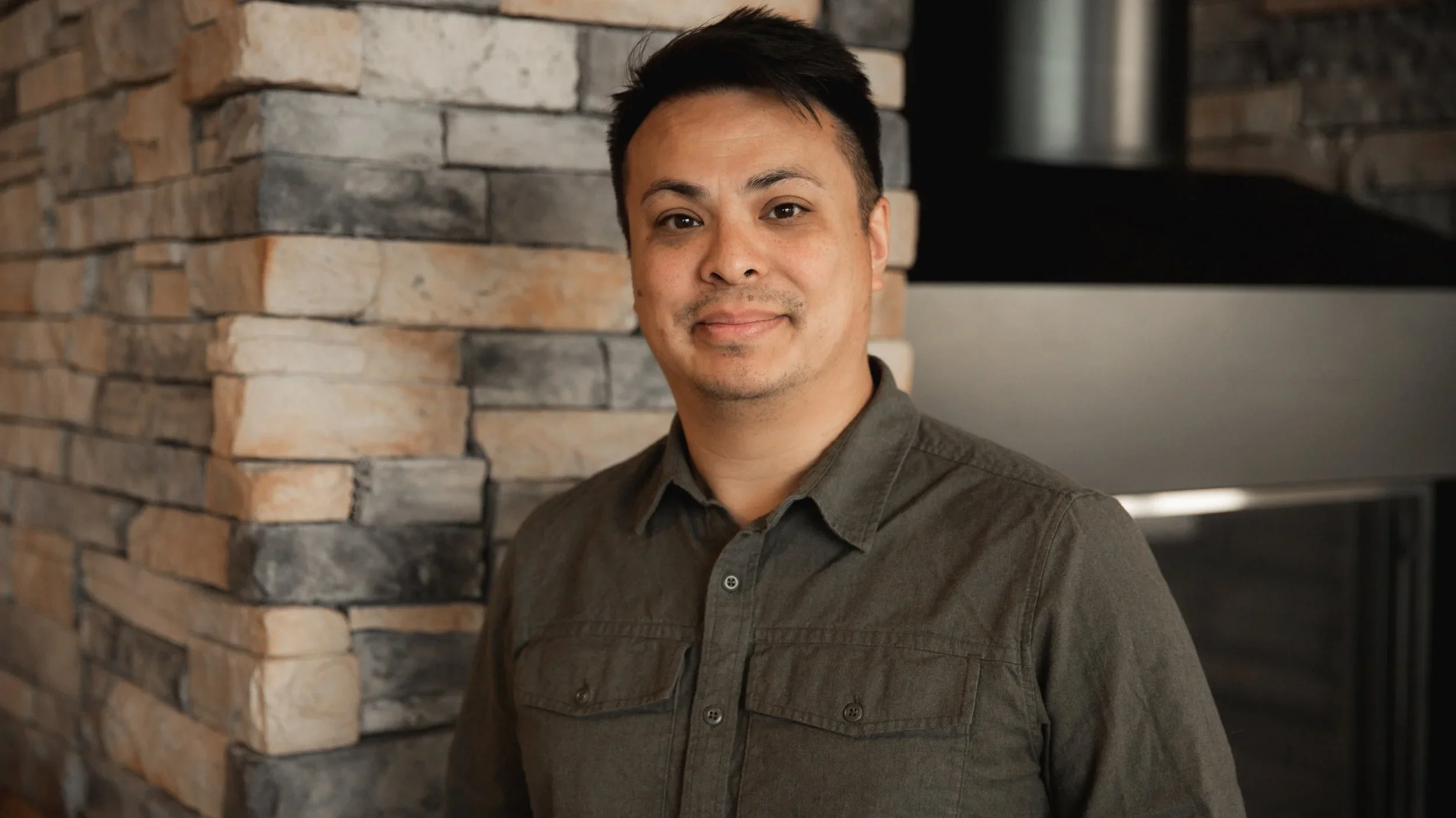 A man with short black hair, wearing a dark green button-up shirt, standing in front of a stone wall with mixed beige and gray stones, smiling slightly.