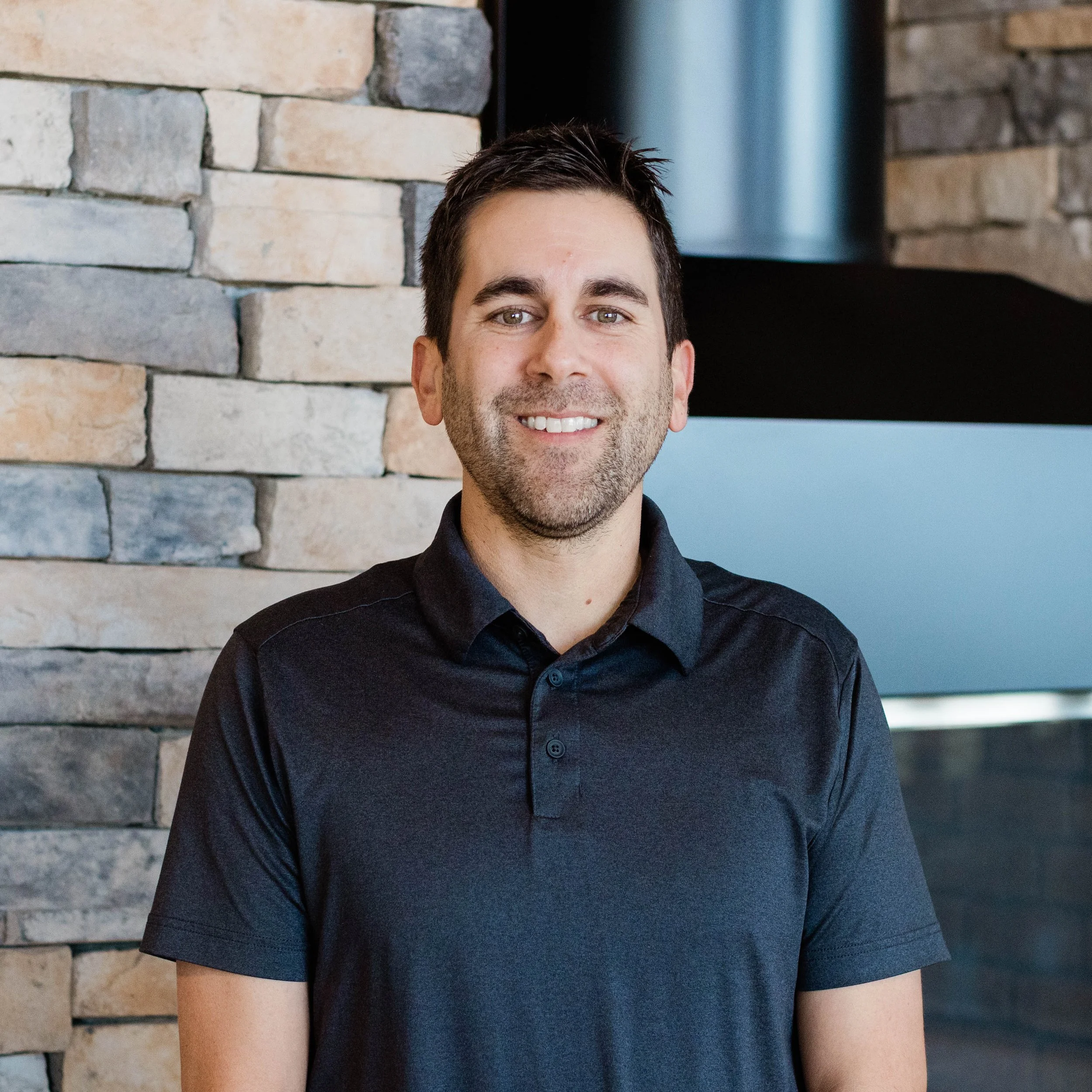 A man with short dark hair and a beard, wearing a dark gray polo shirt, smiling at the camera. He is standing indoors near a brick wall and a fireplace.