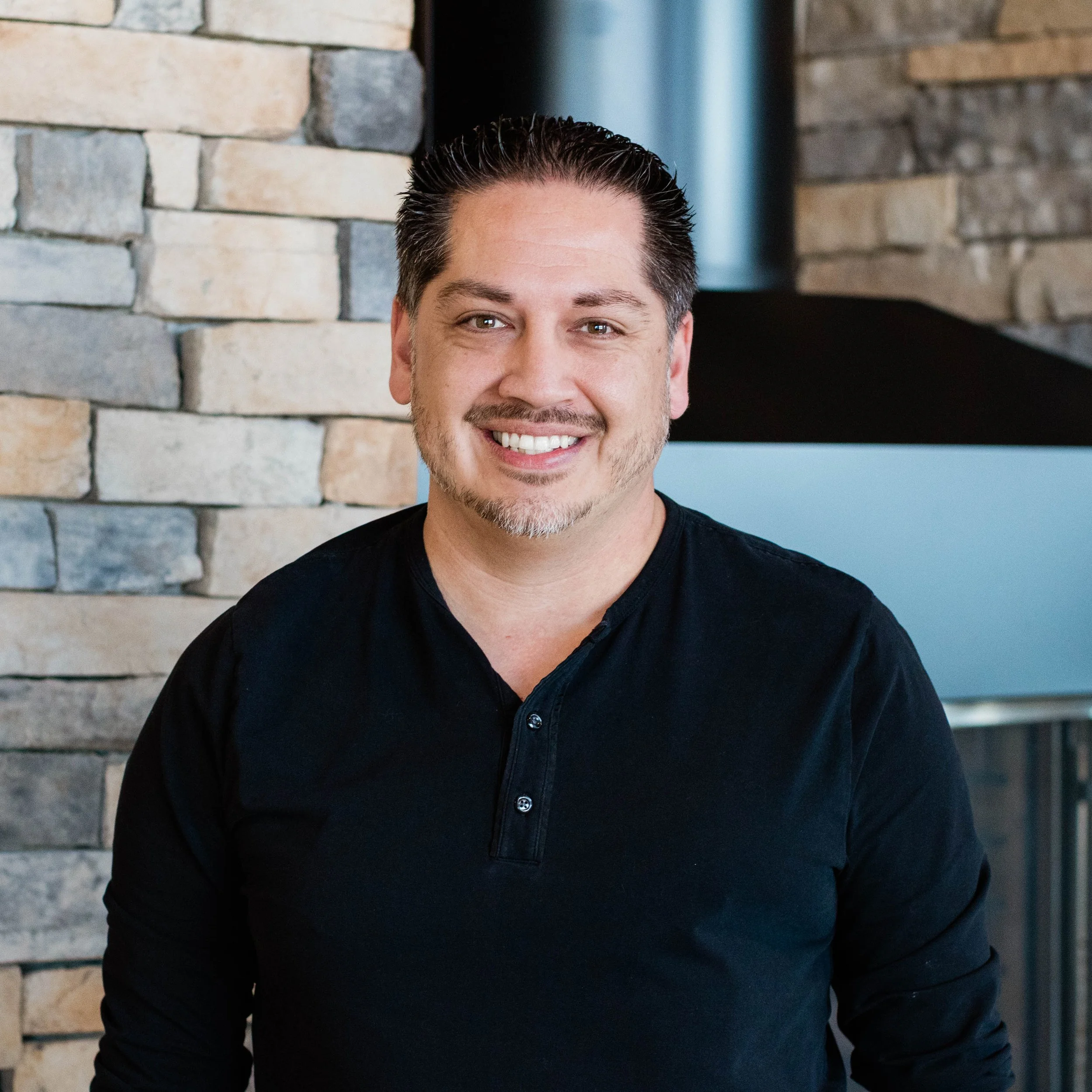 Smiling man in black shirt standing in front of a stone wall and fireplace.