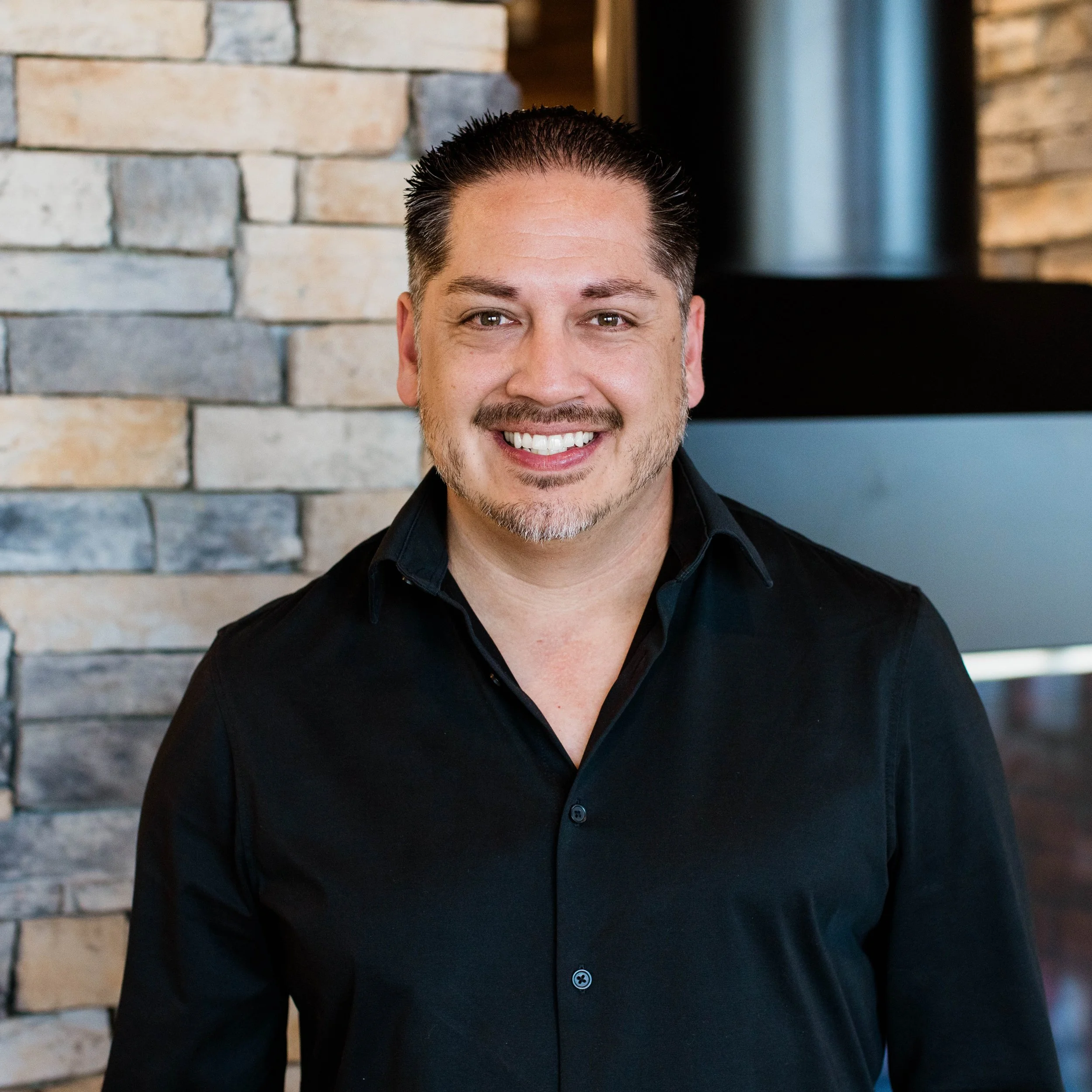 Smiling man in black shirt standing in front of a stone wall and fireplace.