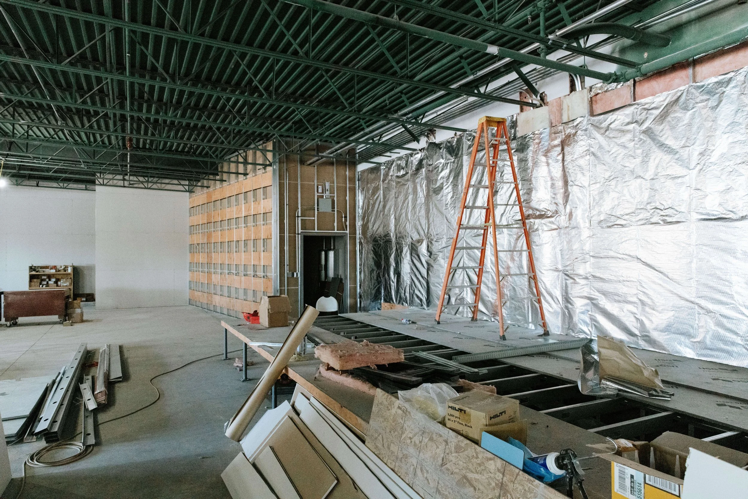 Interior of a construction site with steel framework, an orange ladder, insulated wall, and various construction materials and tools scattered around.