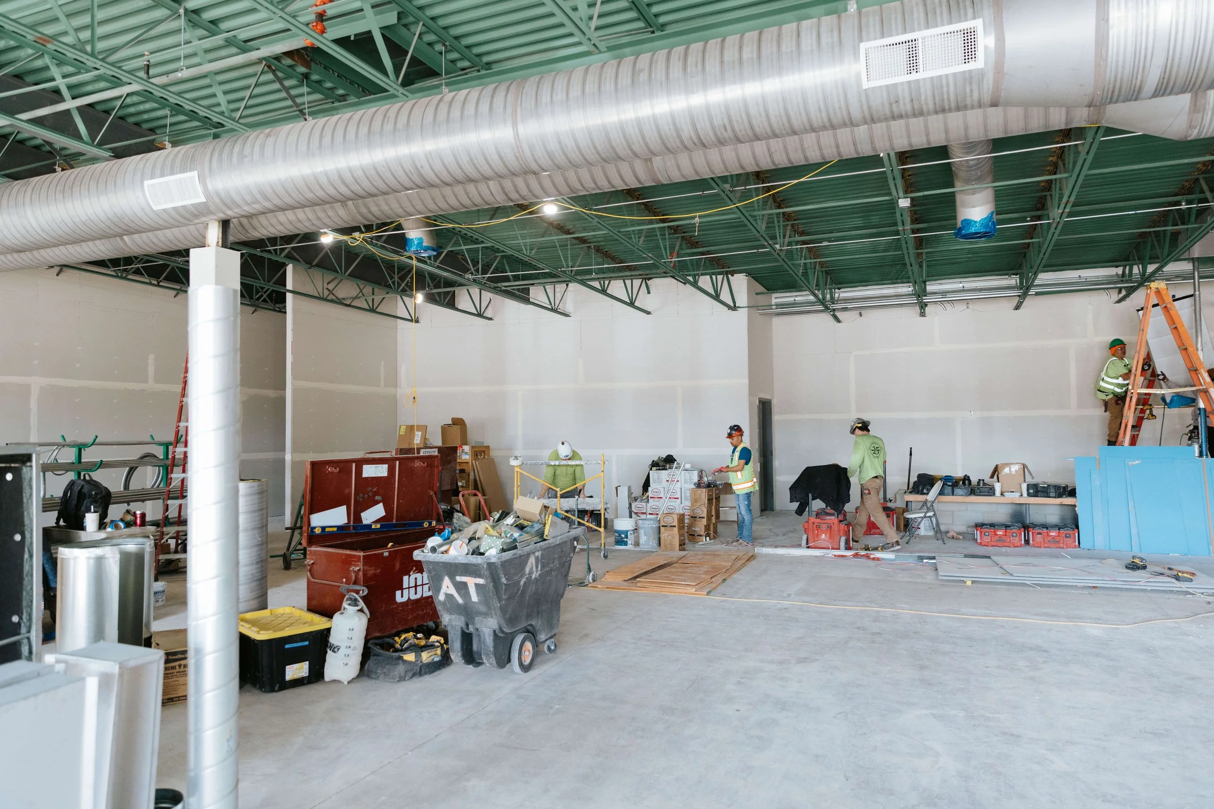Interior view of a building under construction with workers, boxes, tools, and equipment scattered around.