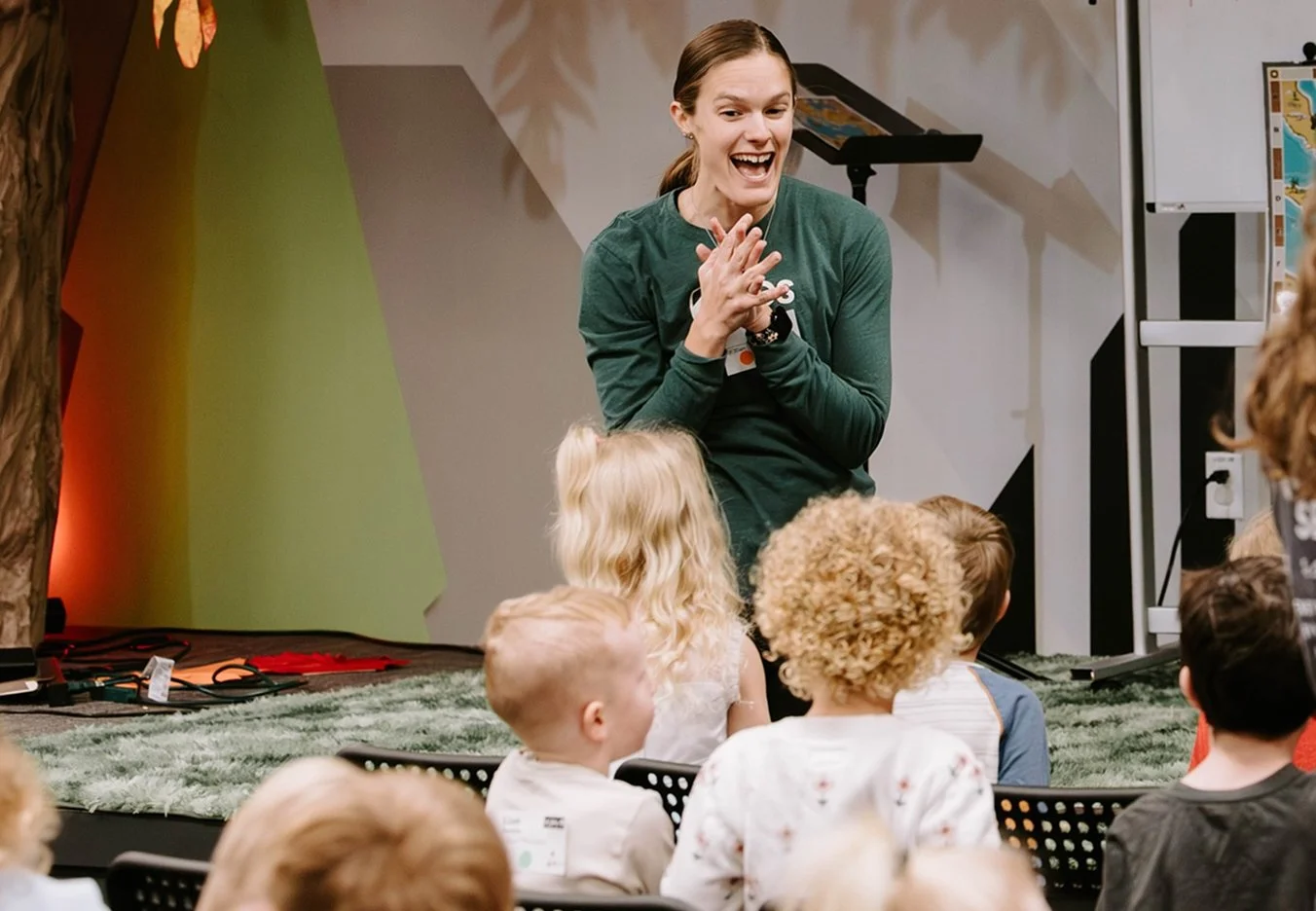 A woman with brown hair, wearing a green long-sleeve shirt, is smiling and gesturing with her hands while speaking to a group of children sitting on the floor during an indoor event.