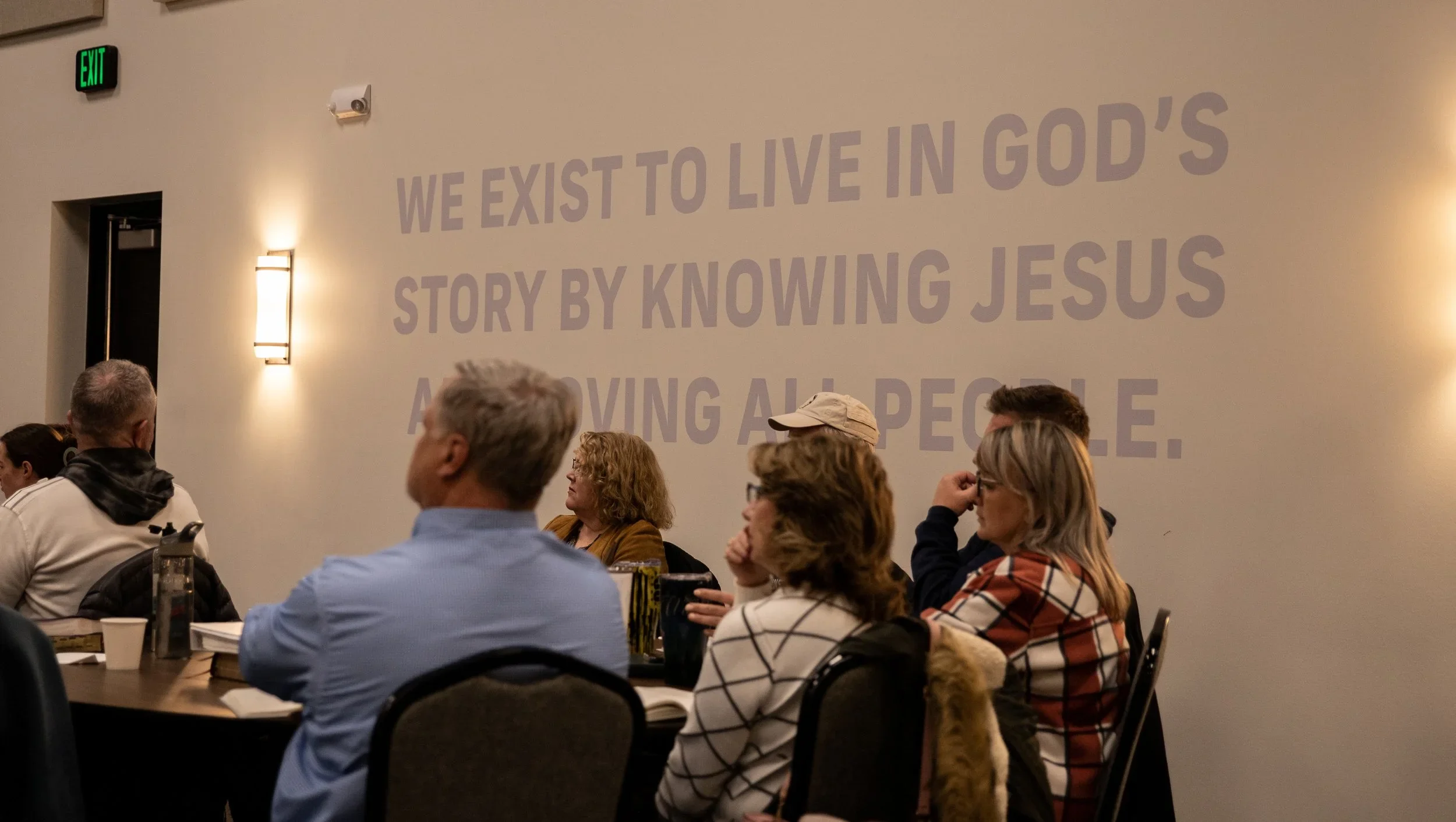 People seated at a table listening during a presentation or meeting in a room with a large wall text that says, 'We exist to live in God's story by knowing Jesus and loving all people.'