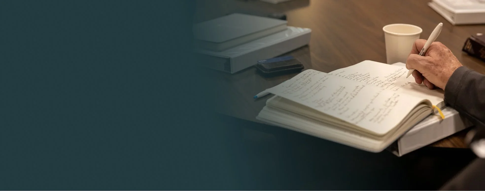 A person writing in a notebook on a desk with a cup, phone, and electronic devices.