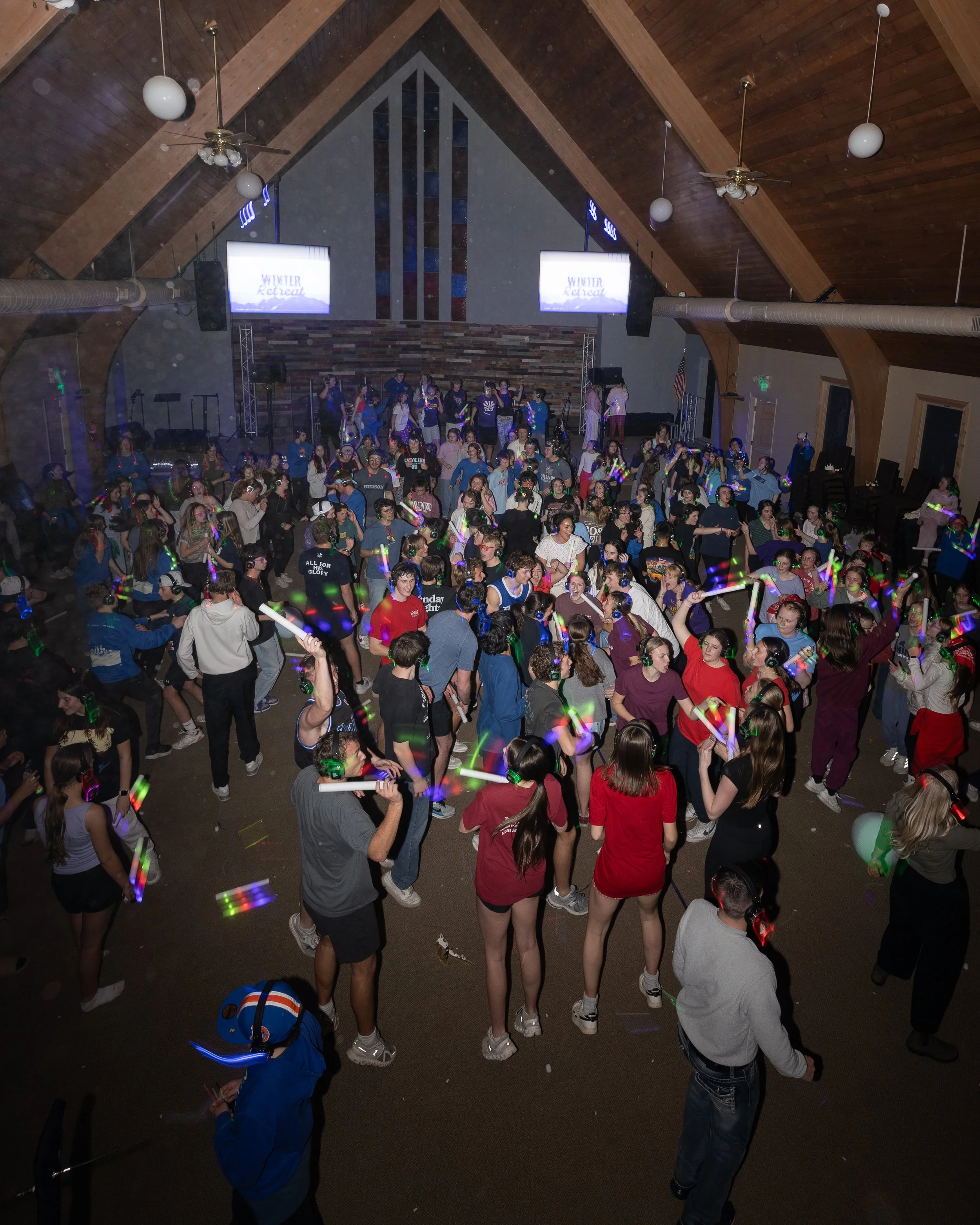 Crowd of young people dancing at a party in a church hall with a stage at the front, decorated with lights and a large patterned wooden wall, during a winter retreat event.