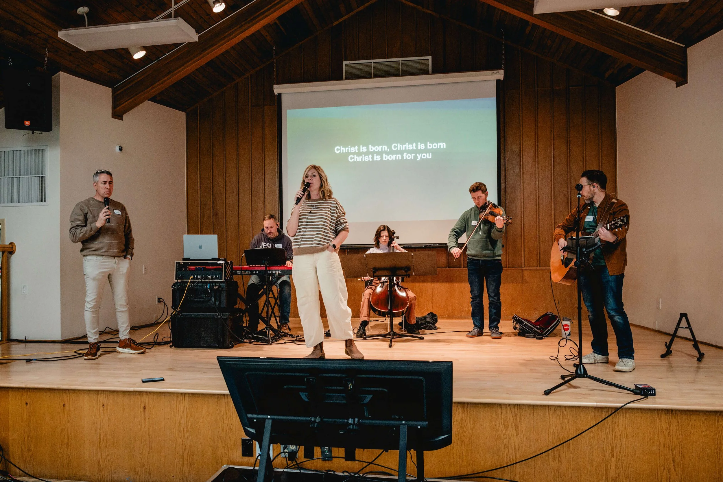 A group of six people on stage performing music and singing during a church service, with lyrics projected on a screen behind them reading 'Christ is born, Christ is born for you'.