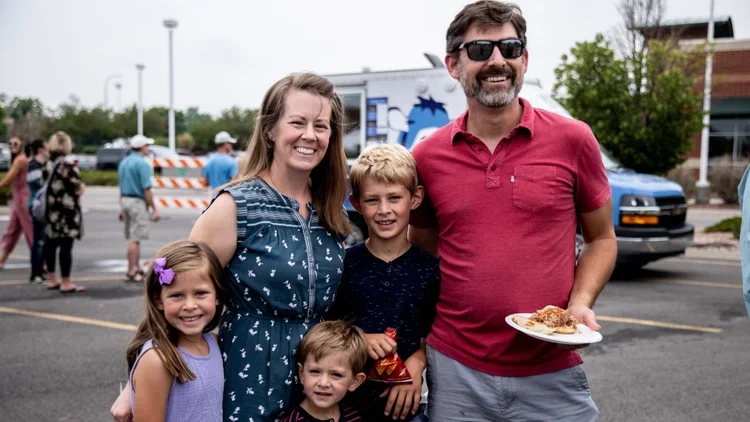 A family of five standing together outdoors in a parking lot, smiling at the camera, with people and a food truck in the background.